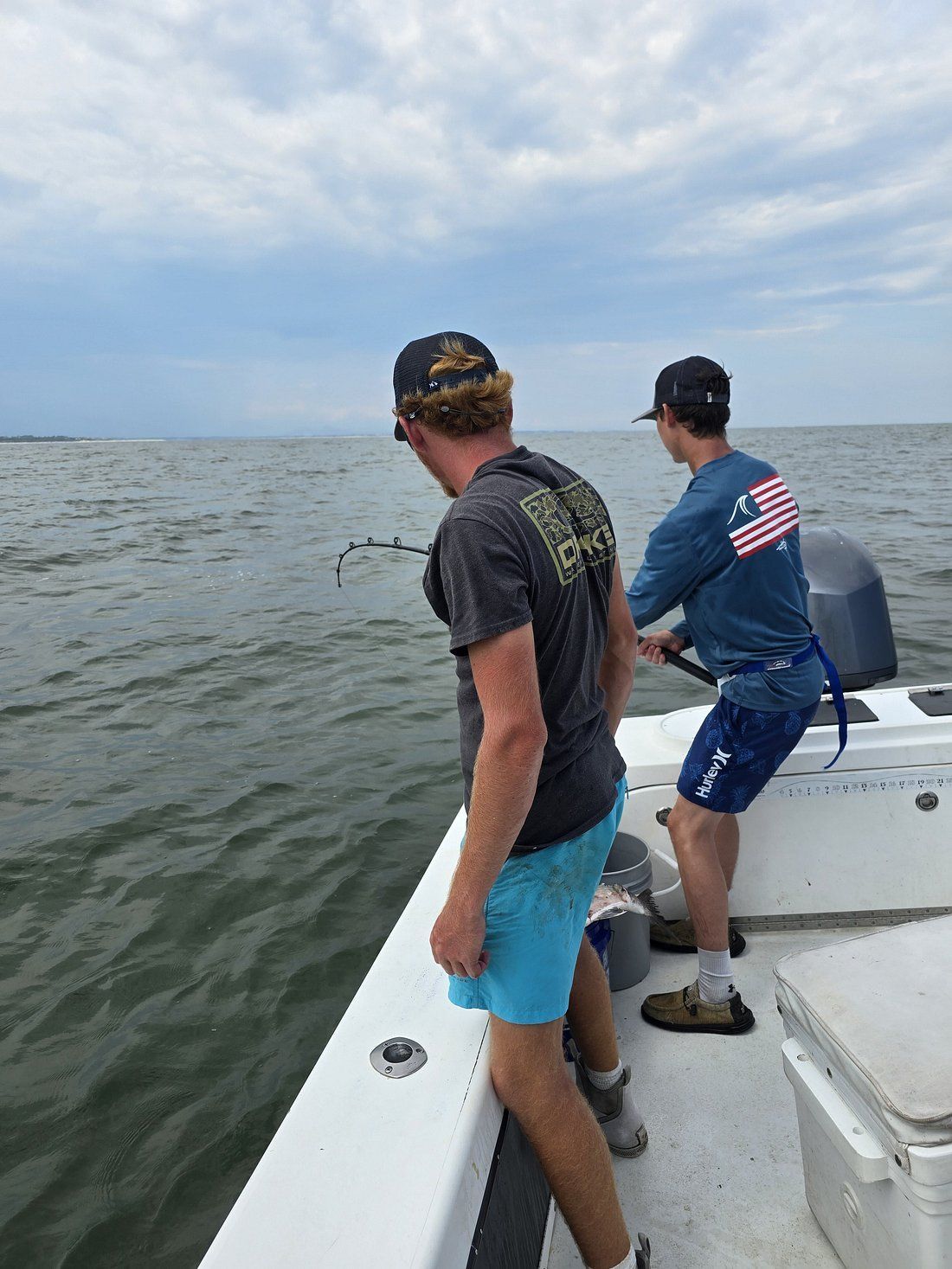 Two men fishing from a boat on a cloudy day; one reeling in, the other watching.
