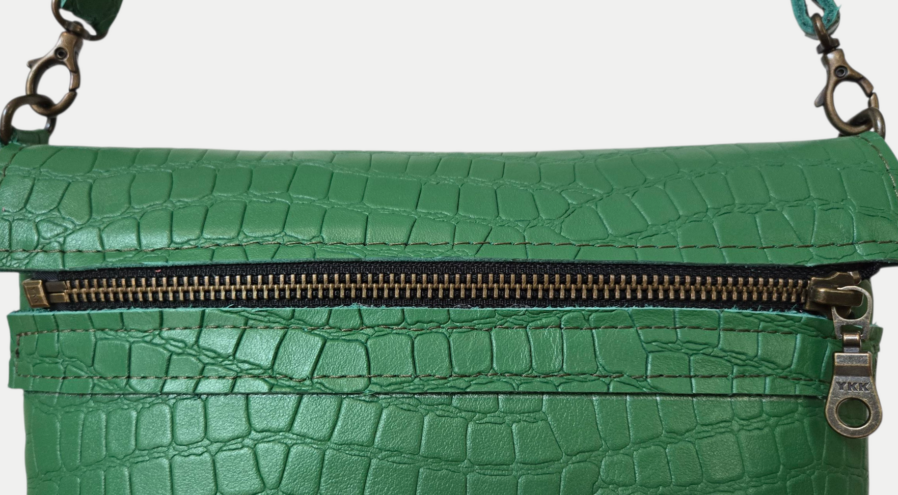 Een close-up van een groene tas van krokodillenleer met een goudkleurige ritssluiting en metalen details.