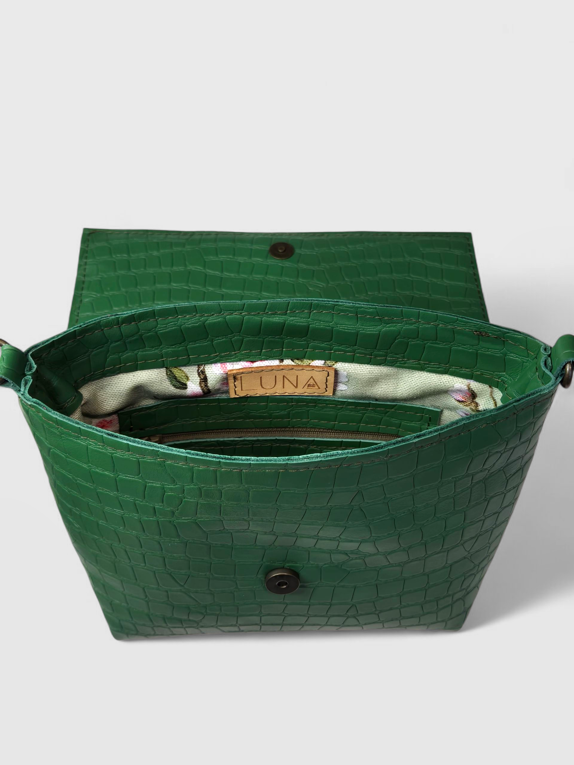 Een crossbodytas met een roze suède klep en een donkergroene body van leer met krokodillenprint, die afsteekt tegen een witte achtergrond.