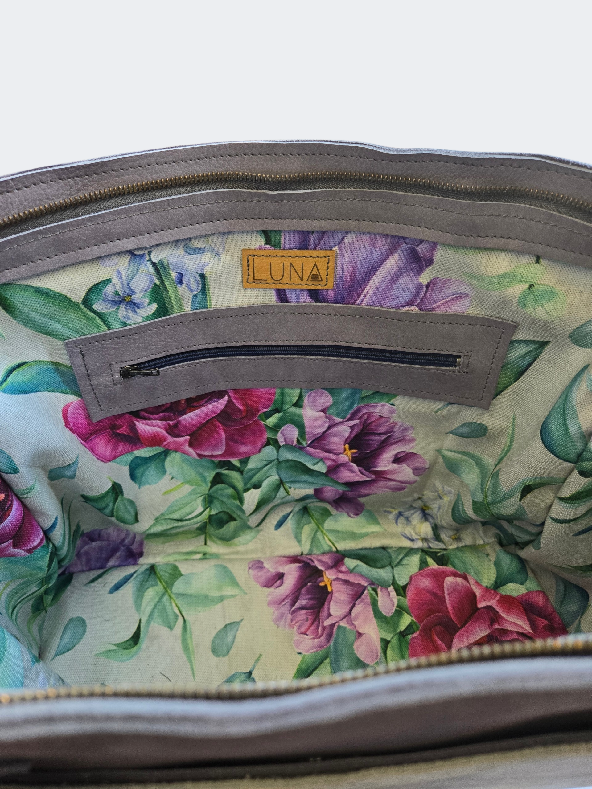 De open binnenkant van een grijze leren tas met een voering van stof met bloemenprint en een klein leren merklabel met de tekst