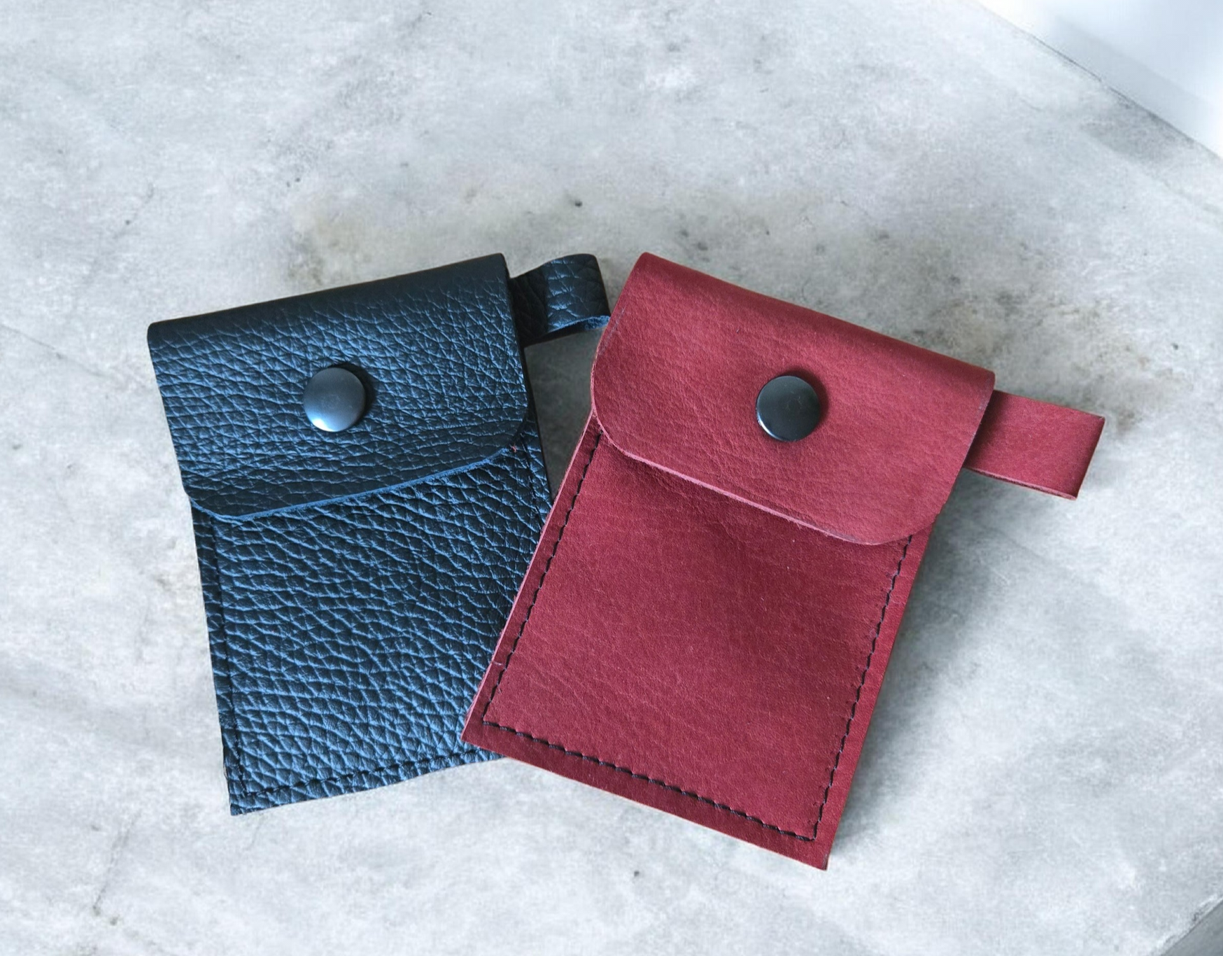 Twee kleine leren buideltjes met drukknoopsluiting, een donkerblauw en een rood, op een lichtgrijze ondergrond.