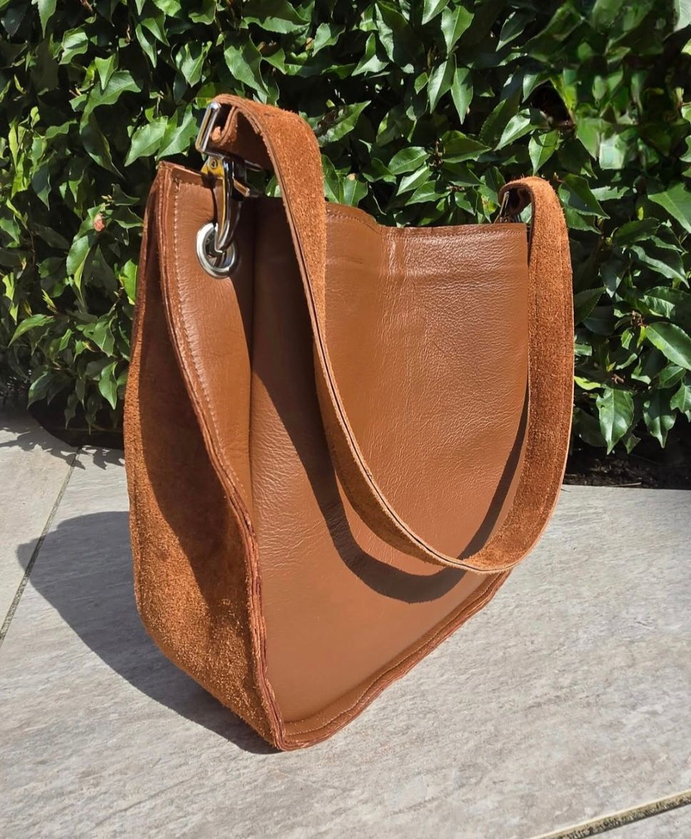 Een bruine leren hobo-schoudertas met zilverkleurige details, geplaatst voor groen gebladerte.