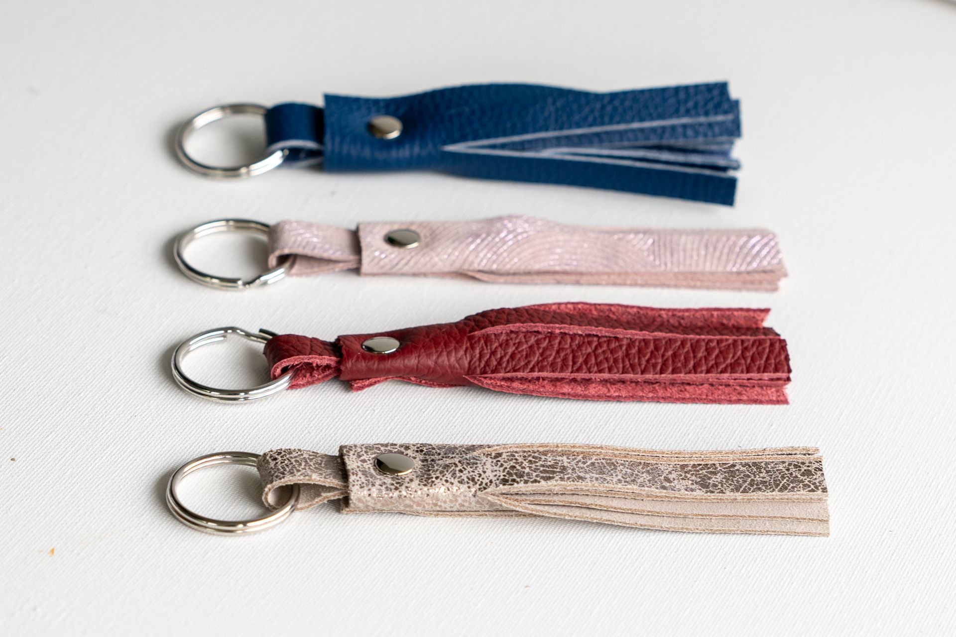 Vier leren sleutelhangers met kwastjes in de kleuren blauw, roze, rood en metallic zilver, verticaal gerangschikt op een witte ondergrond.