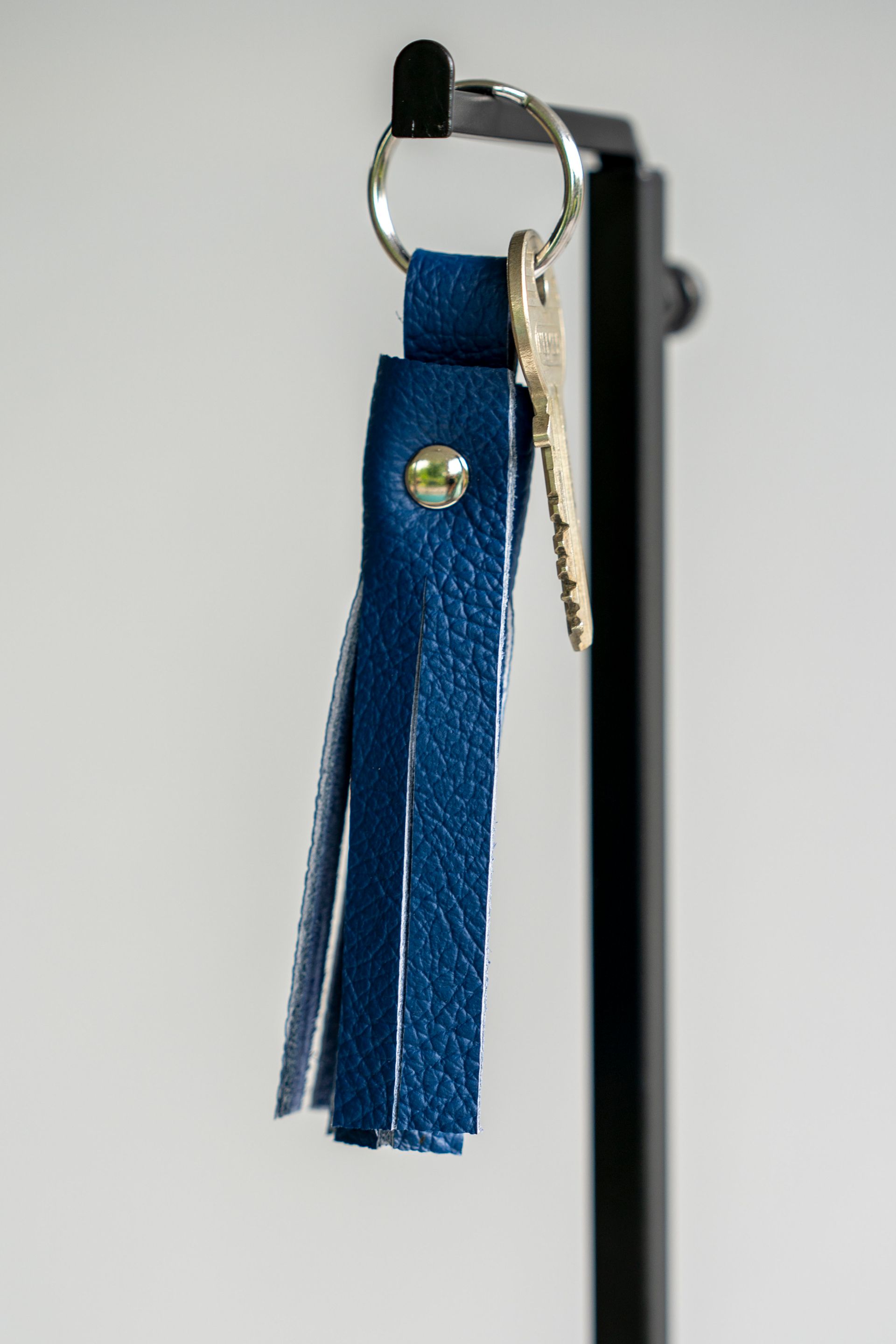 Een sleutelhanger met een blauwe leren kwast hangt aan een zwart metalen displaystandaard, met een metalen sleutel aan de ring.