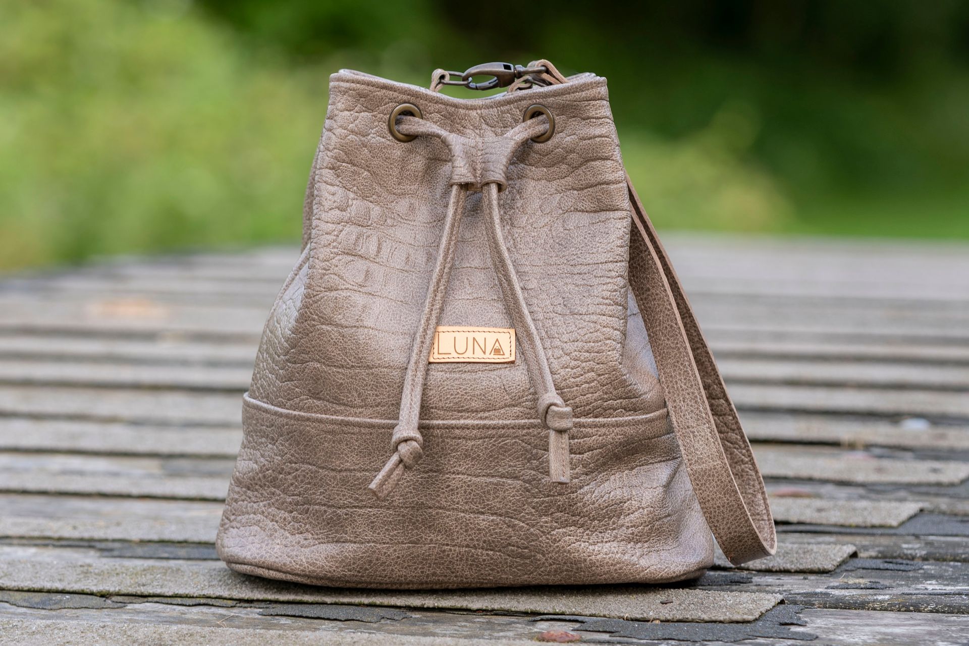 Een gestructureerde, metallic bronzen bucket bag met trekkoord en schouderband, die op een houten ondergrond staat.