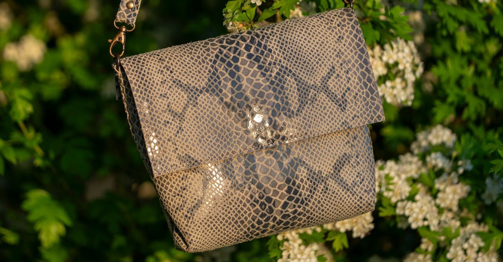 Een schoudertas met slangenprint hangt tegen een achtergrond van groene bladeren en witte bloeiende struiken.