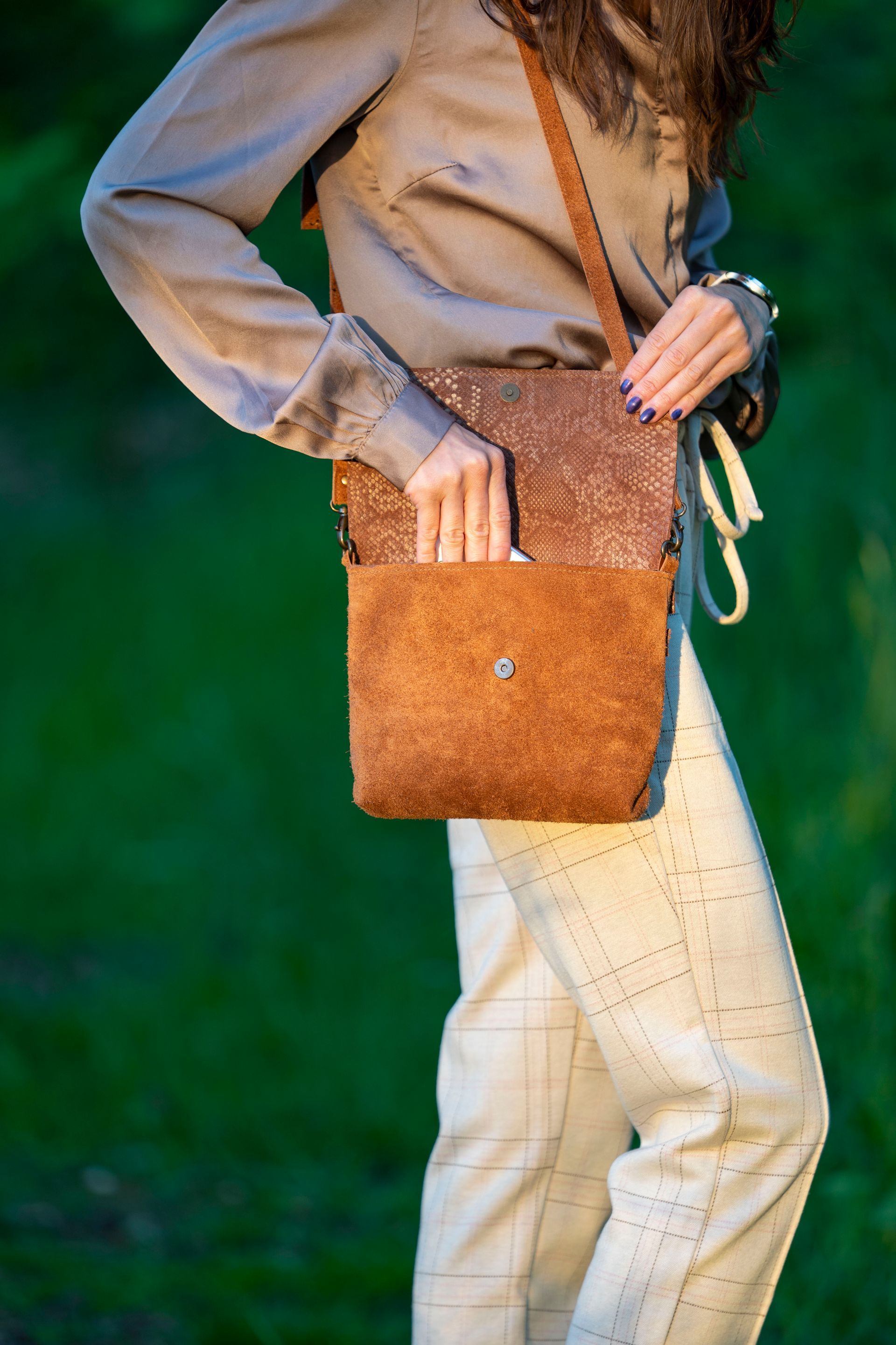 Een persoon in een beige shirt en een lichtgekleurde broek met patroon draagt een bruine leren schoudertas met structuur in een grasveld buiten.