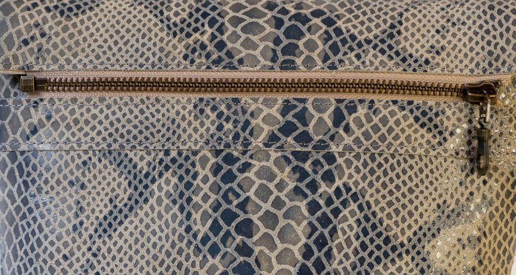 Een close-up van een ritsvakje op een beige en grijze tas van stof met slangenprint.