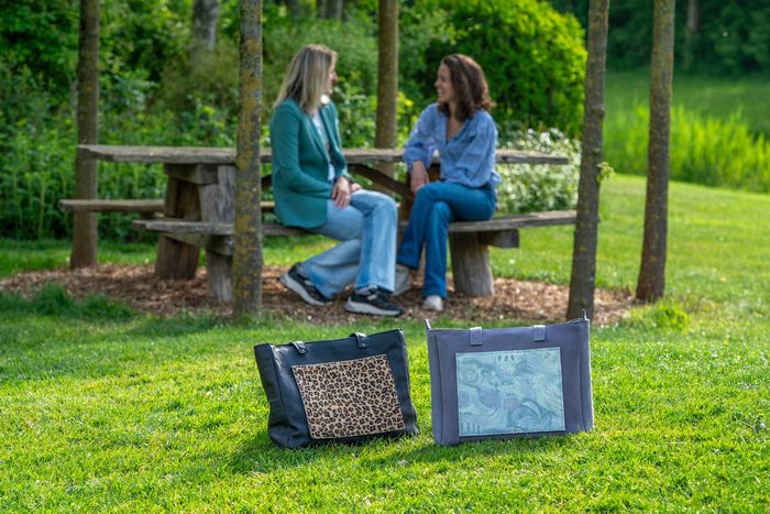 Twee mensen zitten te praten aan een picknicktafel in een park, naast twee draagtassen op het gras.