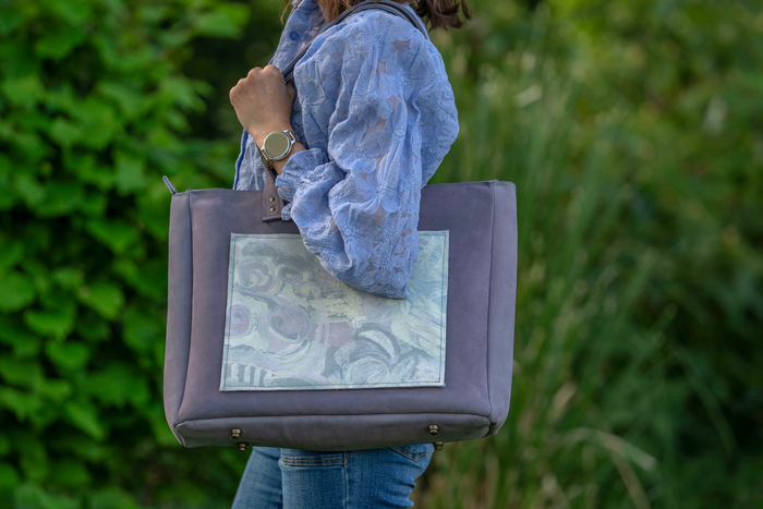 Persoon in blauwe trui met laptop in de hand, lachend buiten voor groen gebladerte.