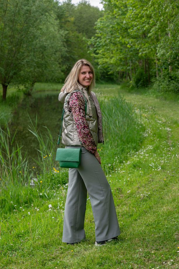 Vrouw staat op een graspad in een groen bosrijk gebied, gekleed in een grijze broek en een jas met een patroon, en heeft een groene tas bij zich.
