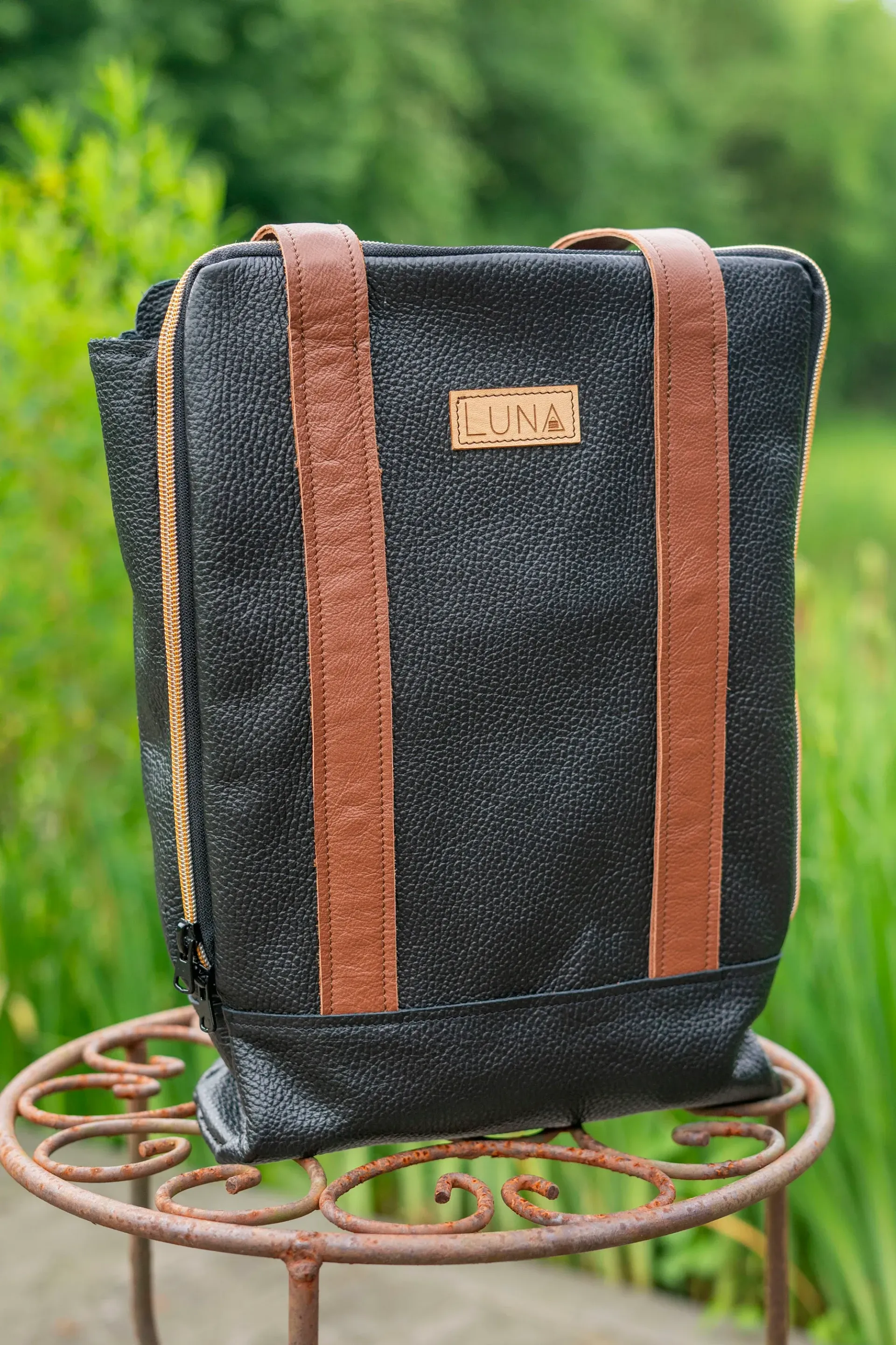 Een zwarte, glinsterende tas met twee verticale, bruine leren bandjes en een klein merklabel, die op een verroeste metalen tuinstandaard staat.