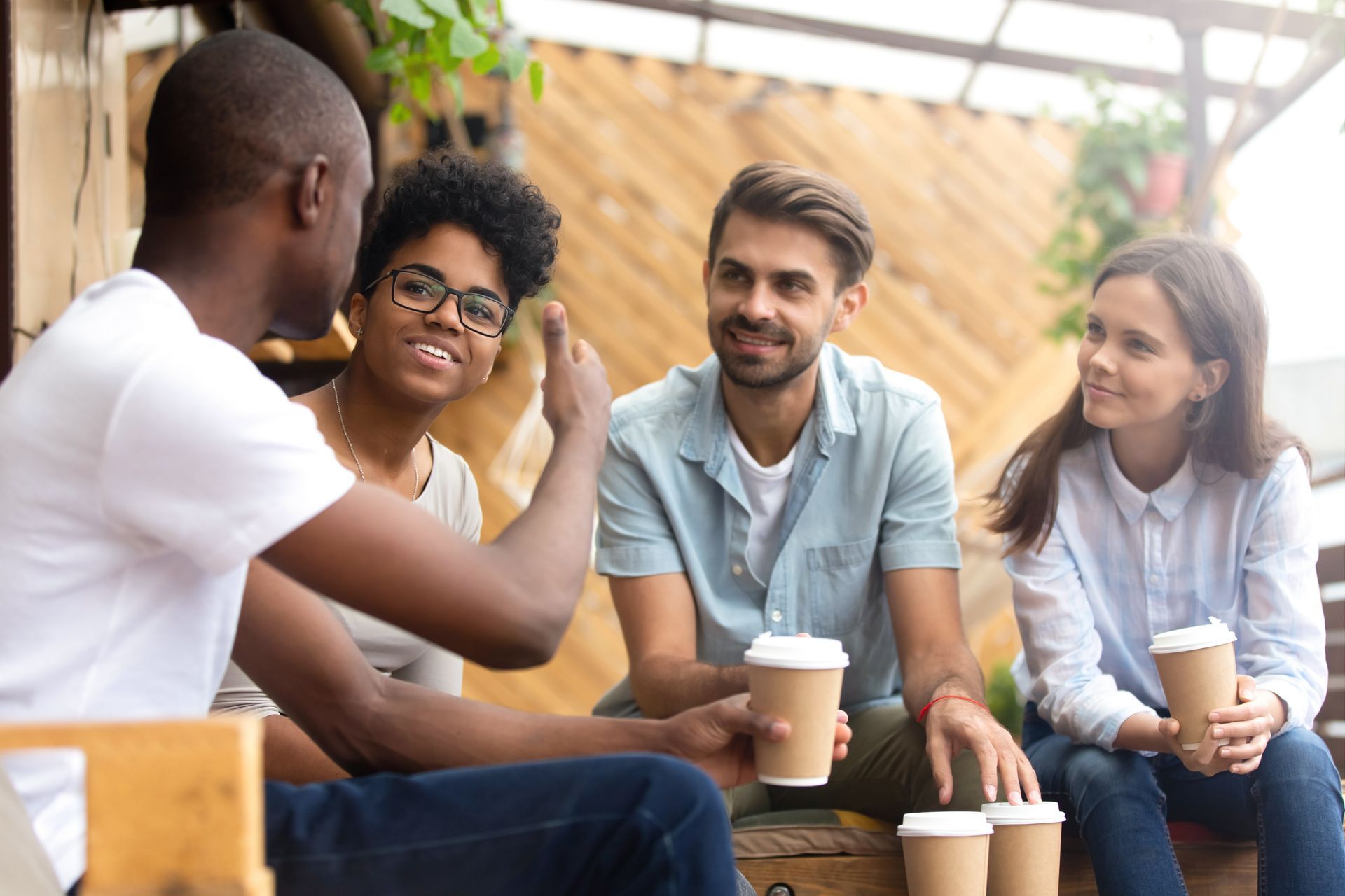 Four people in a cafe, talking and holding coffee cups. The setting is outdoors.