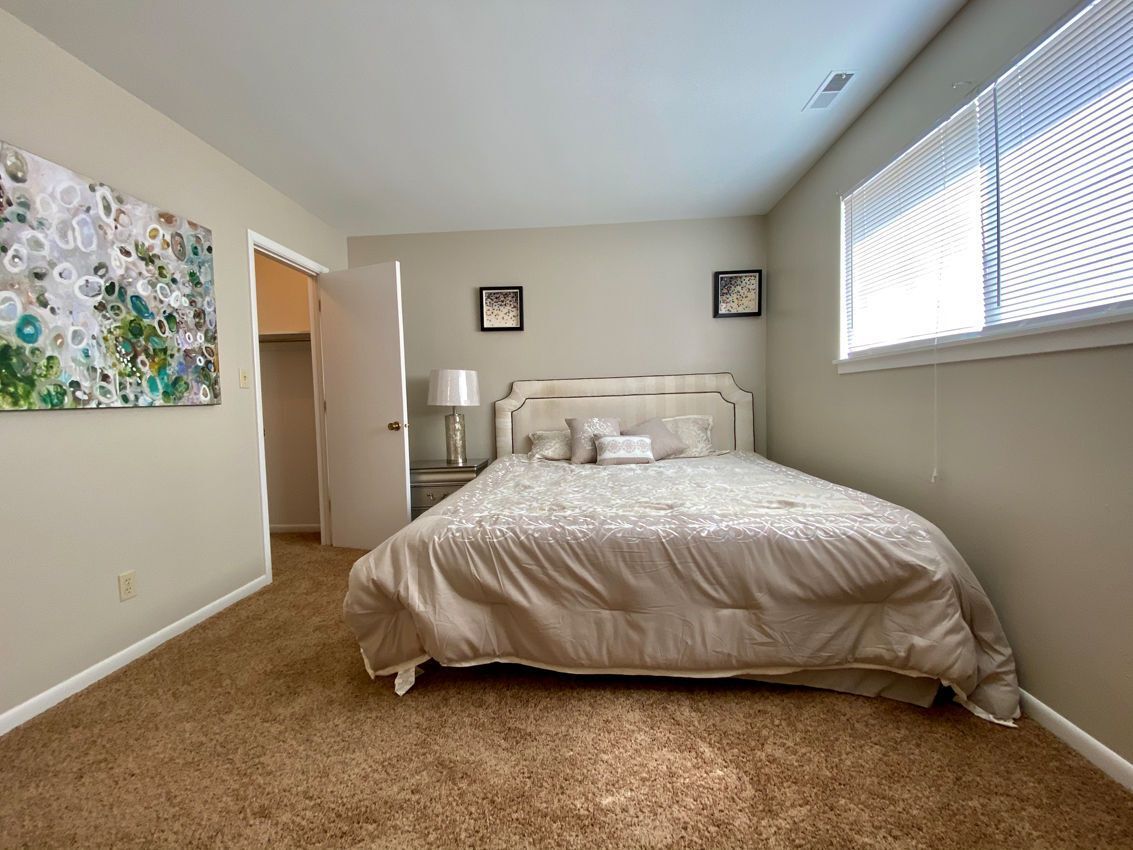 Bedroom with beige walls, bed with tan comforter, artwork, window with blinds.