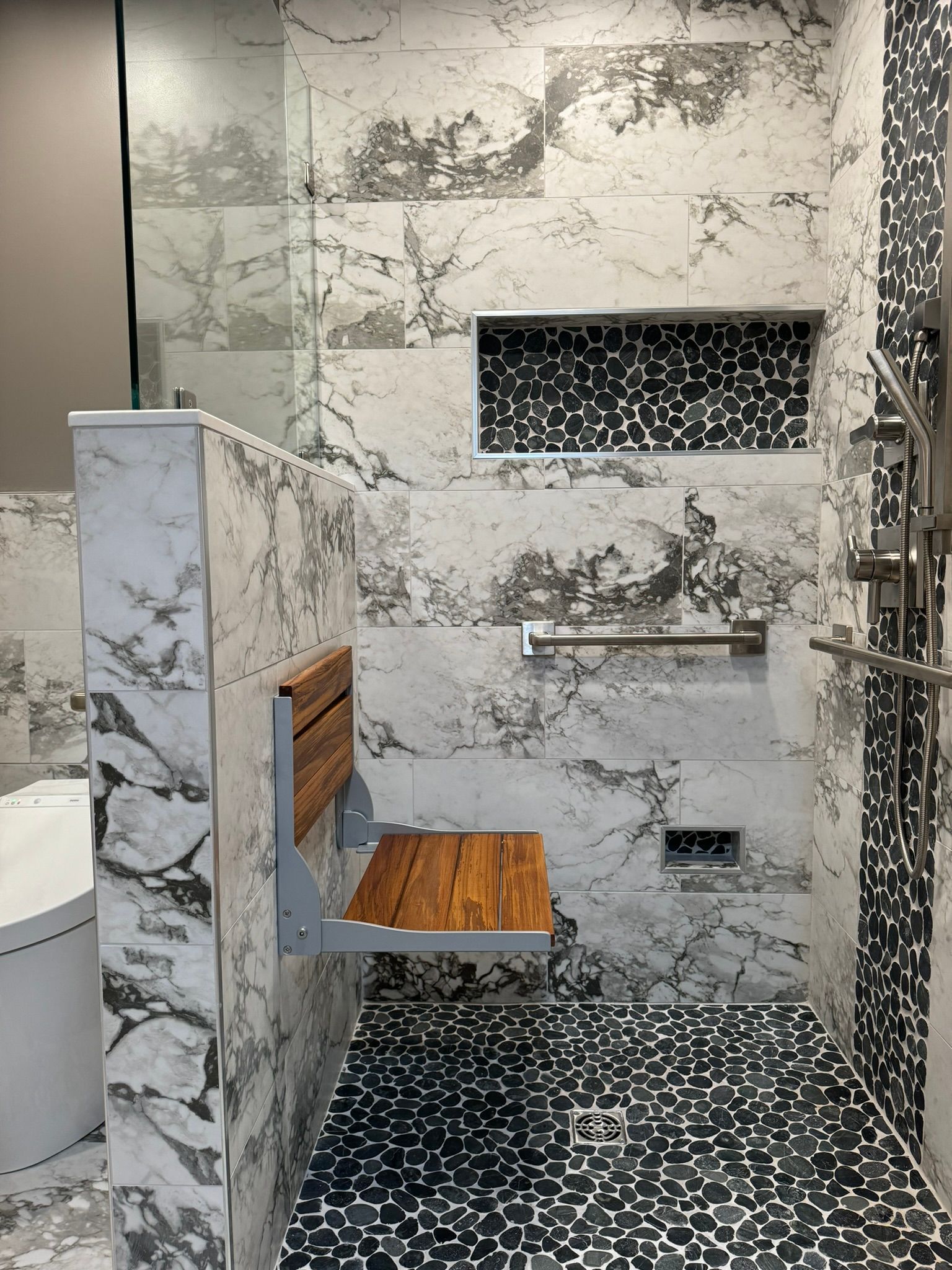 Marble-tiled bathroom with built-in wood bench, black patterned floor, and recessed wall shelf.