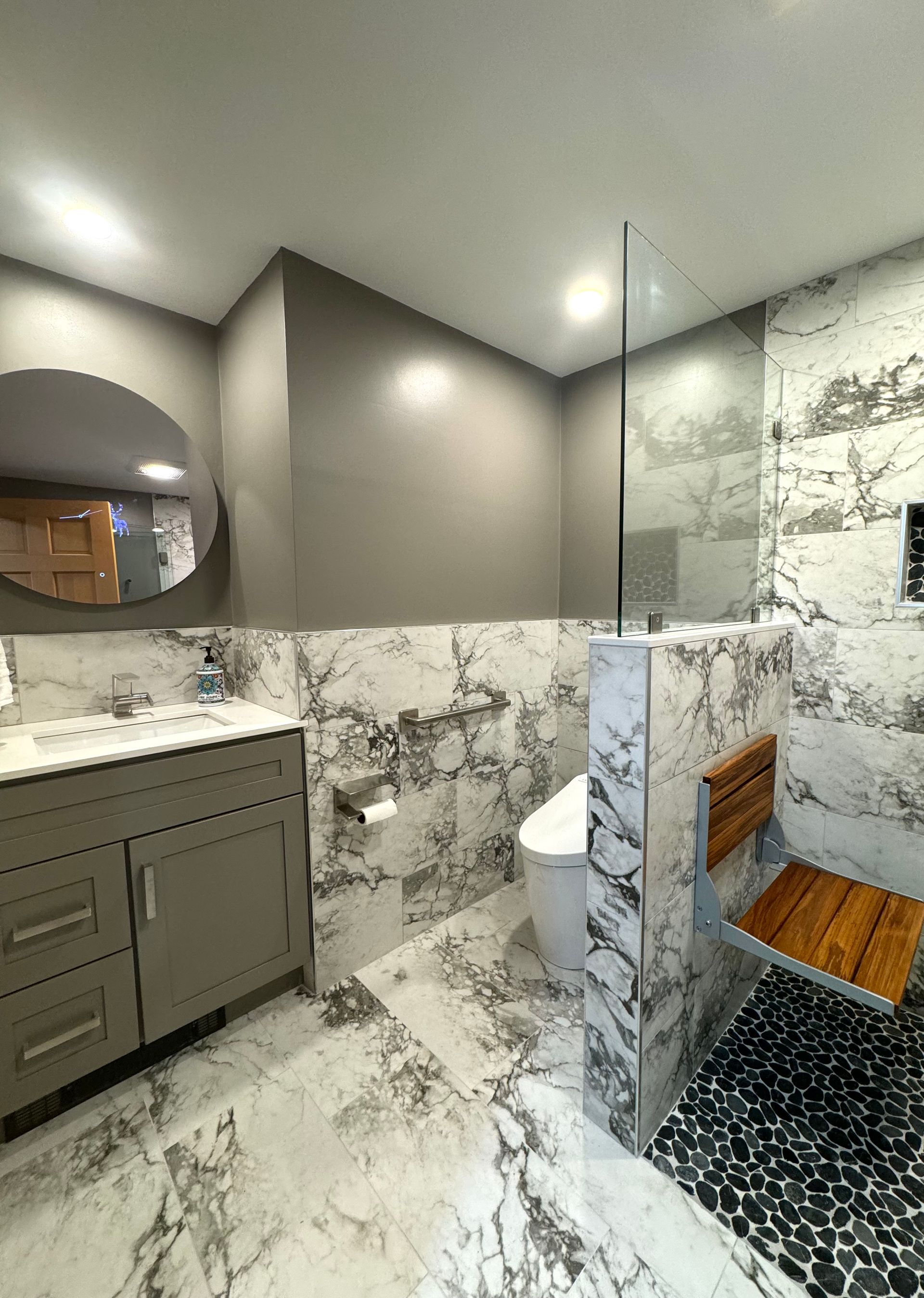 Marble-tiled bathroom with gray vanity, round mirror, glass shower, and black mosaic floor.