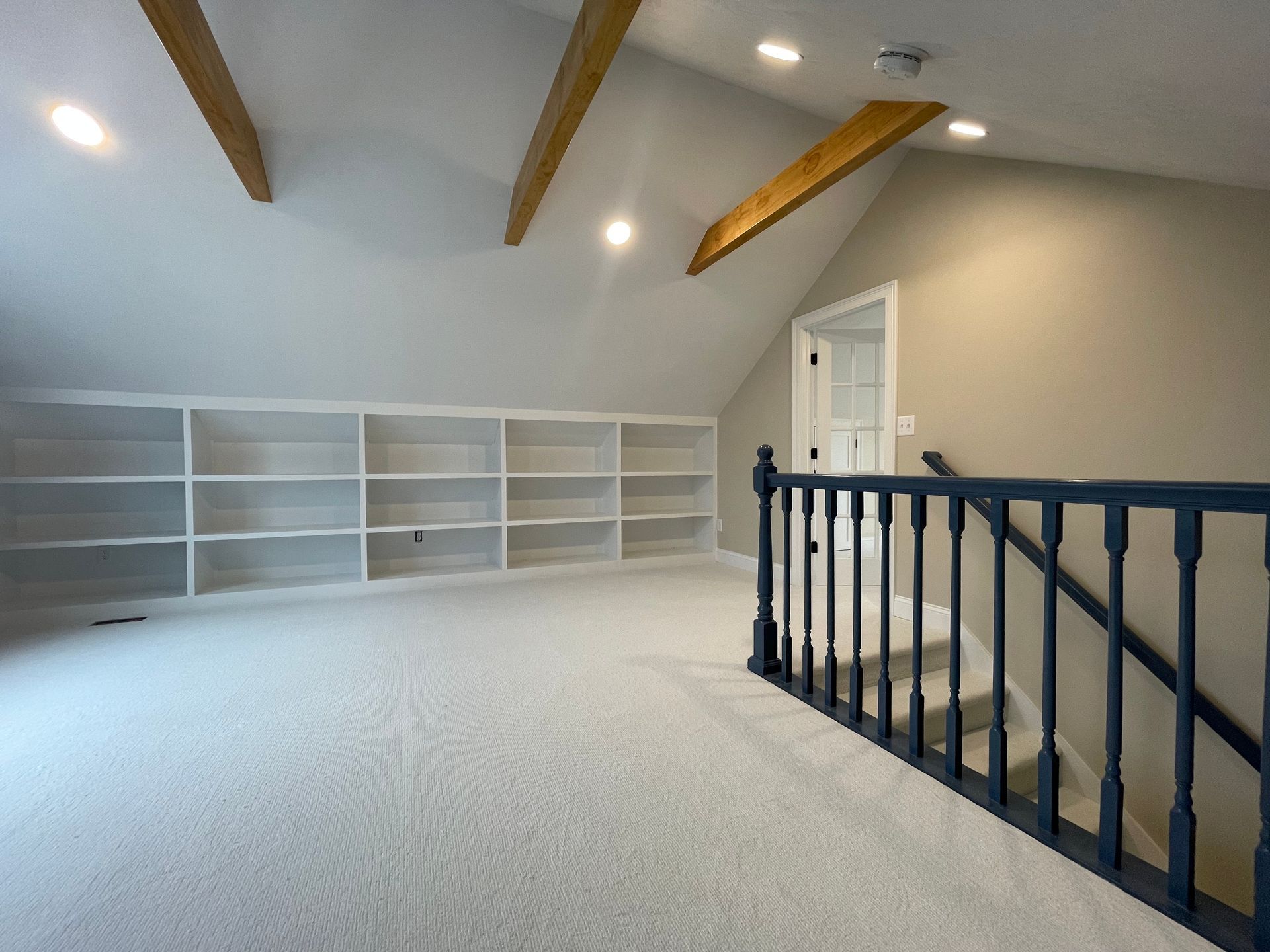 Spacious attic room with white built-in shelves, exposed wooden beams, and a black stair railing