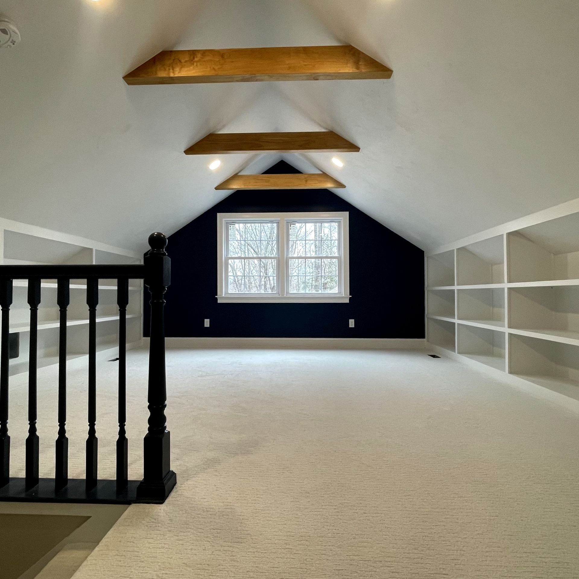 Empty attic room with vaulted white ceiling, exposed beams, navy accent wall, and central window.
