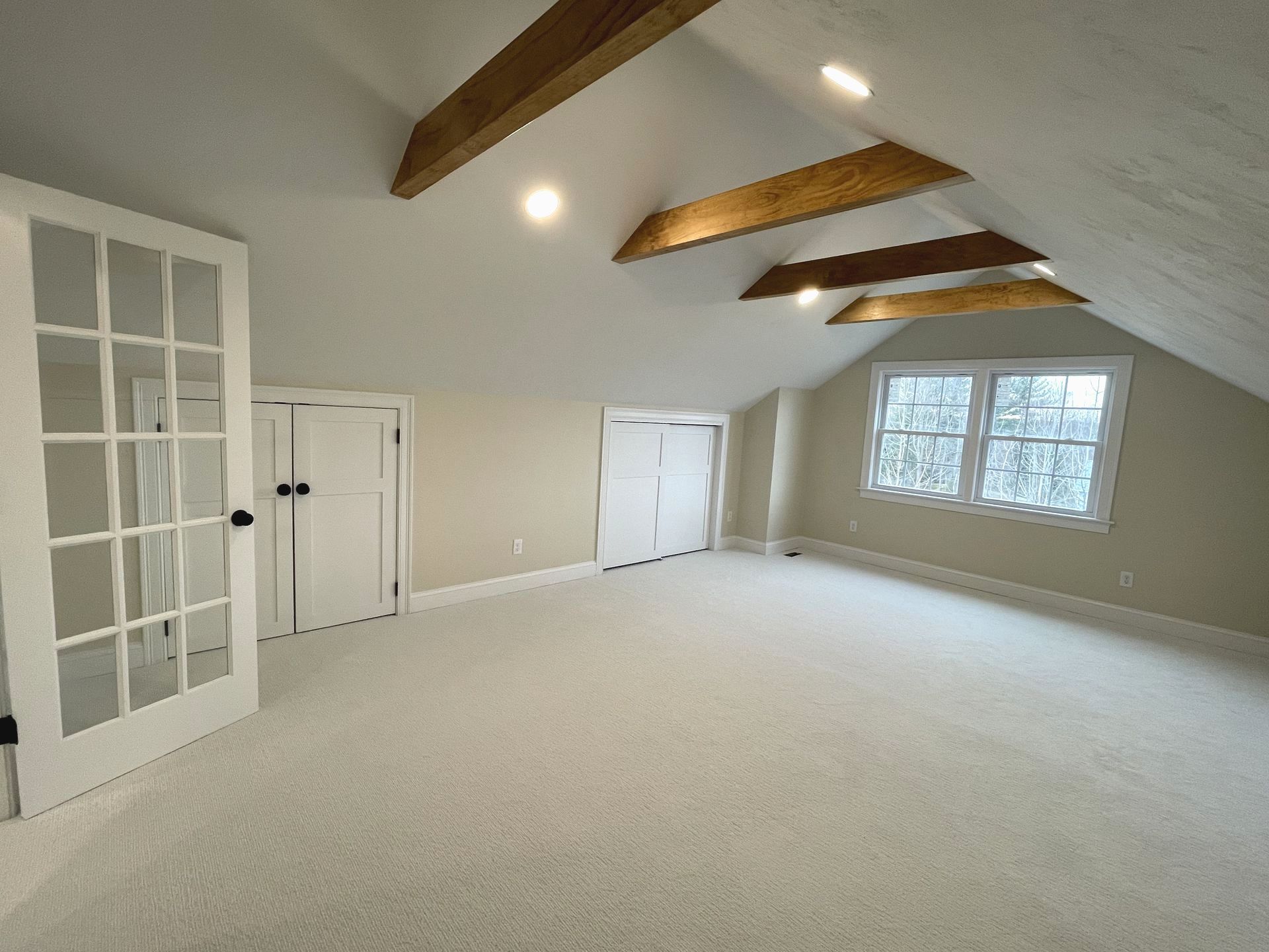 Empty beige attic room with vaulted ceiling, exposed beams, French doors, and a window seat area