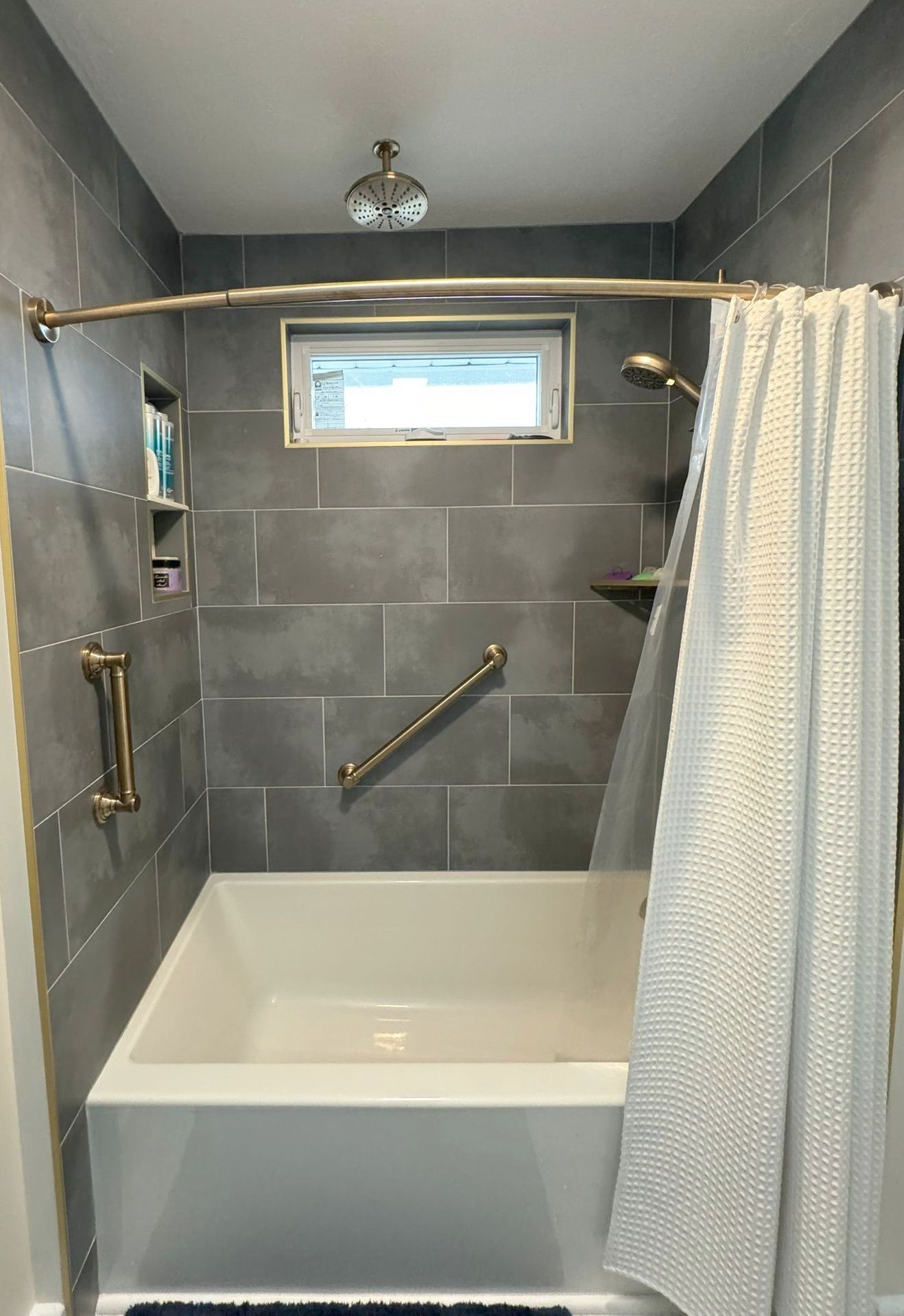 Small gray-tiled shower with tub, gold grab bars, shower curtain, and a frosted window.