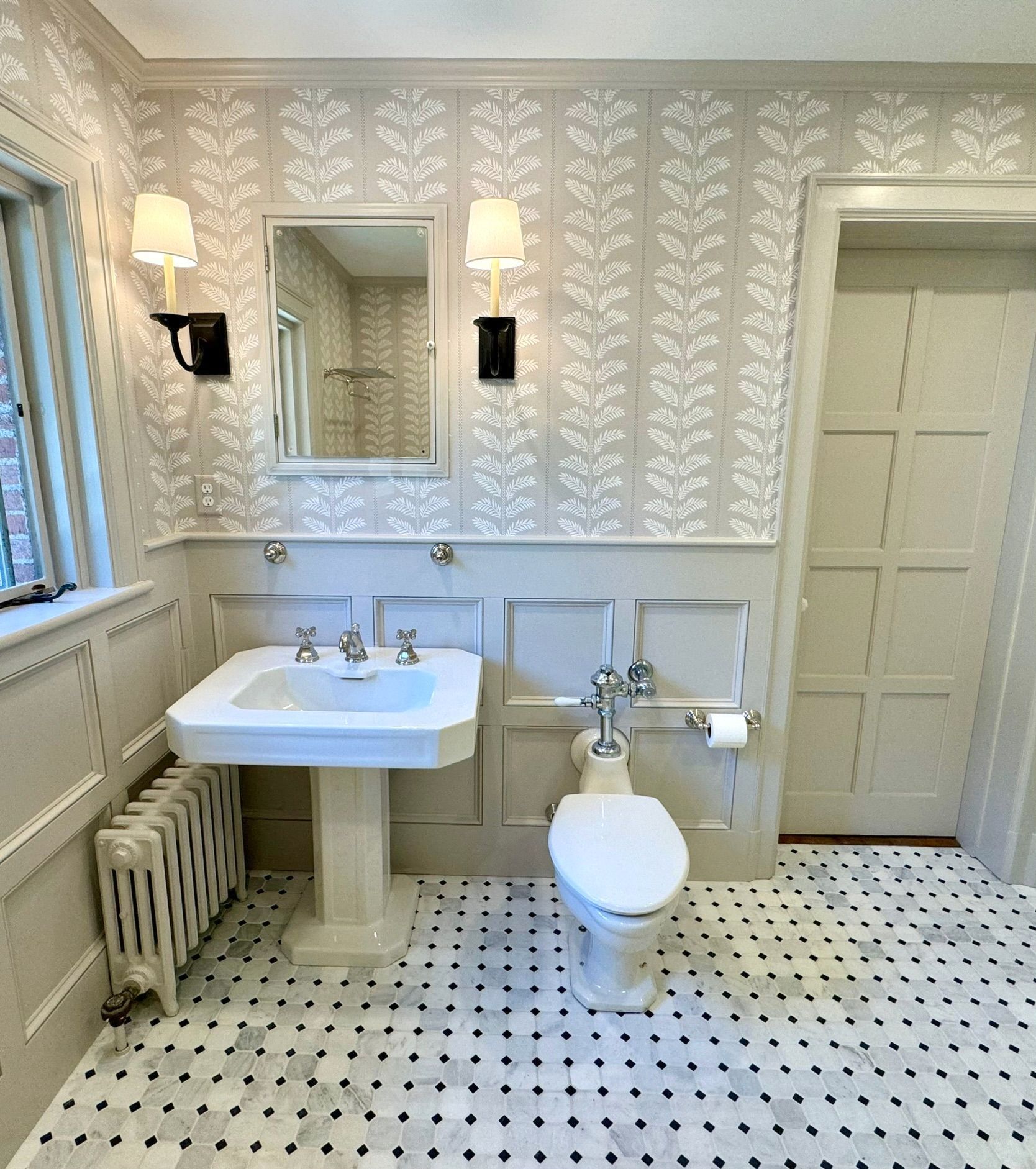 Vintage bathroom with pedestal sink, toilet, wall sconces, mirror, and black-and-white tiled floor.