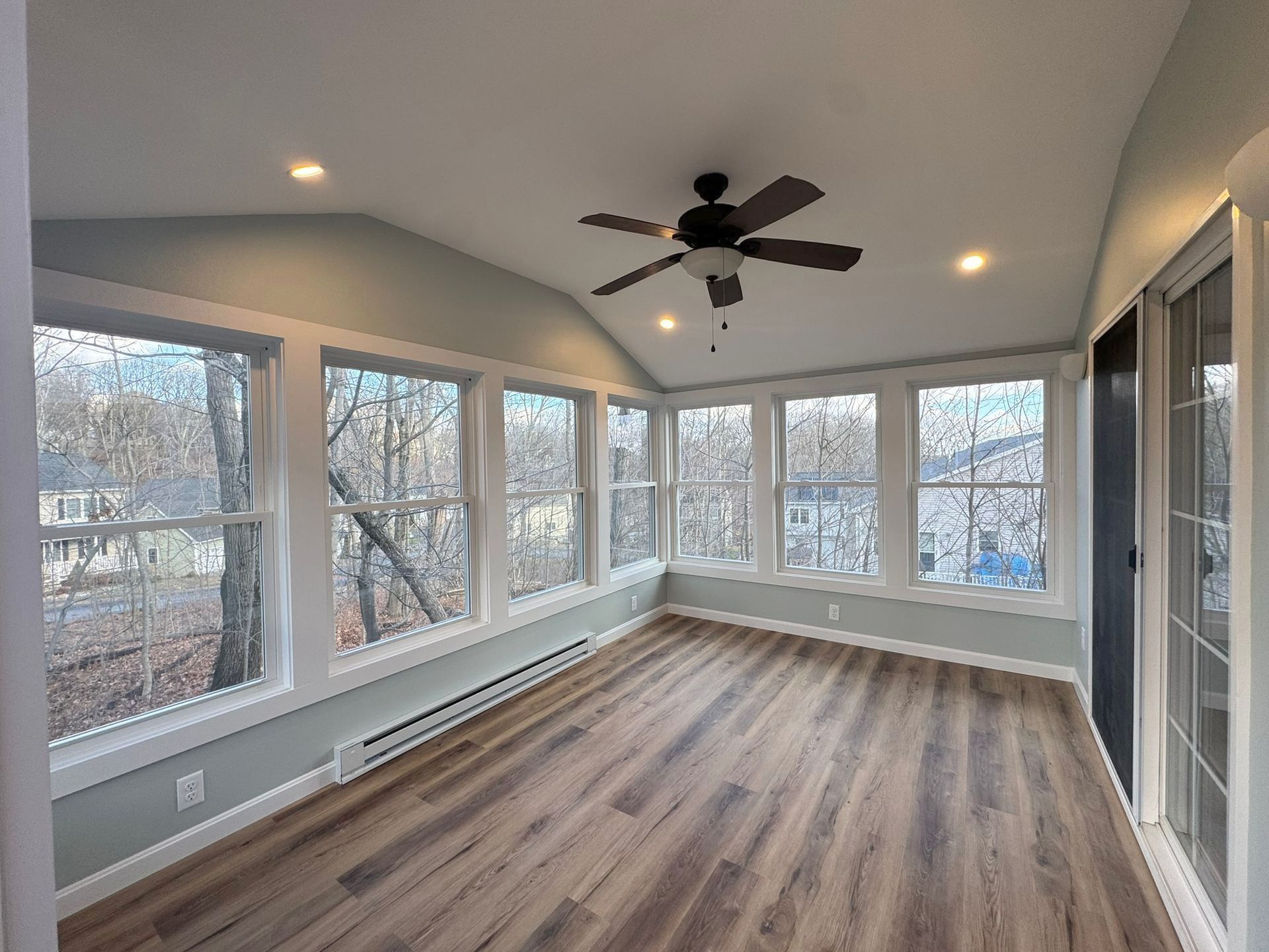 Bright sunroom with large windows, wood flooring, and ceiling fan overlooking trees