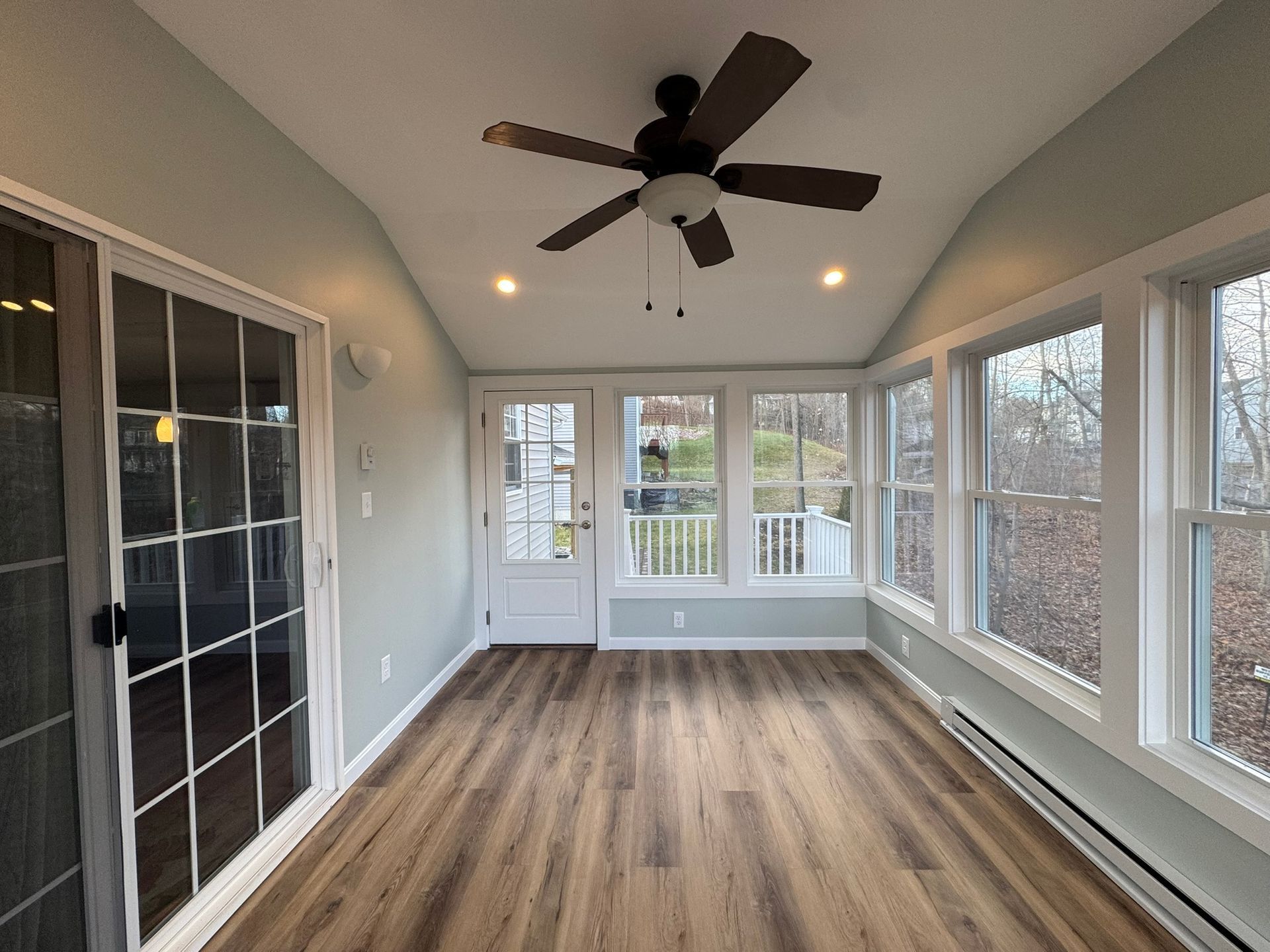 Bright sunroom with wood floors, ceiling fan, white walls, and large windows overlooking greenery.