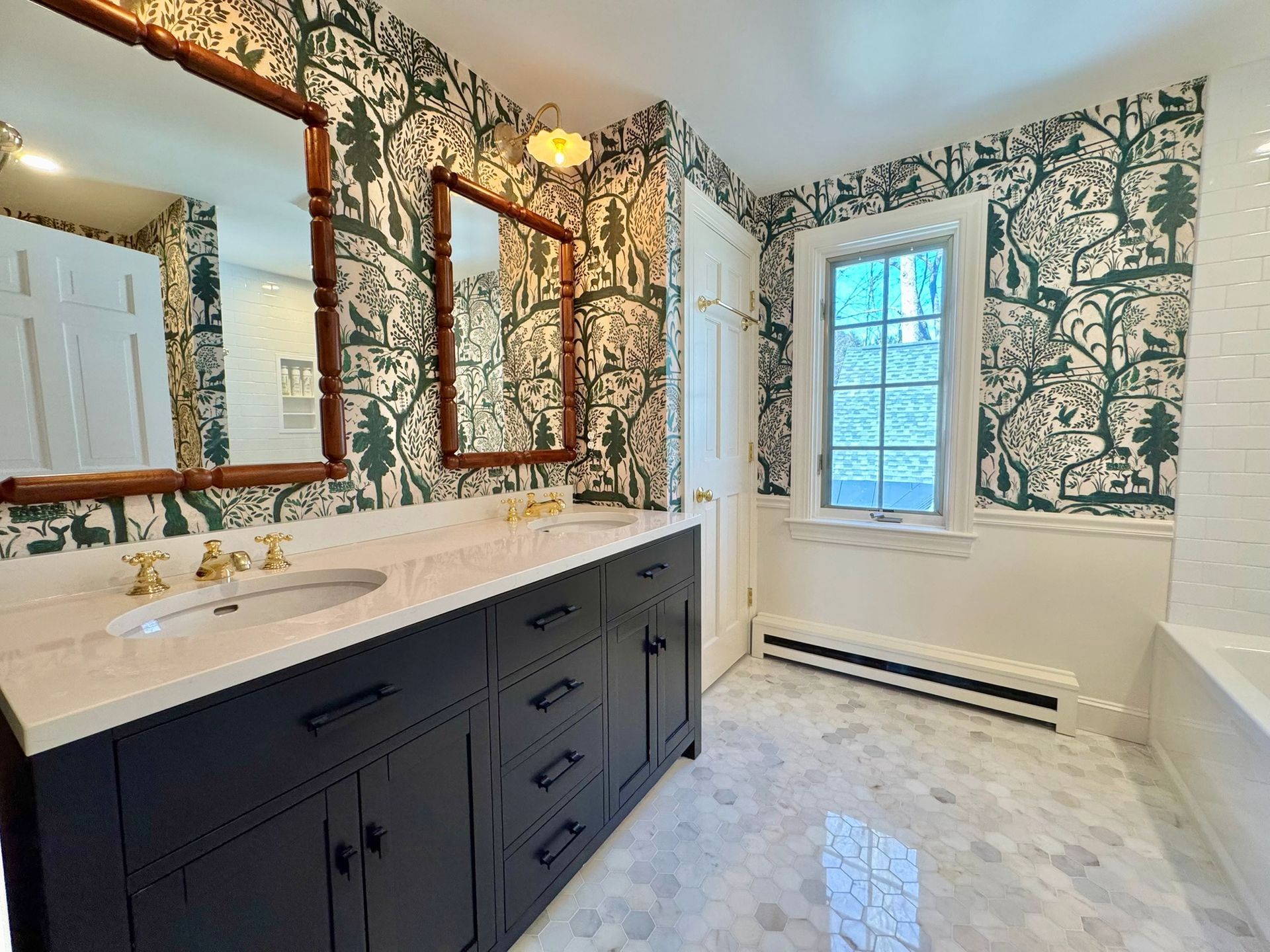Modern bathroom with dark vanity, white counters, floral wallpaper, large mirror, and window.