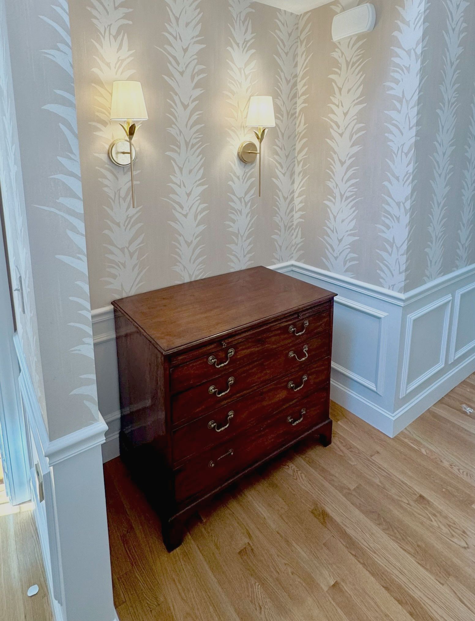 Wooden dresser in a softly lit corner with cream patterned wallpaper and wall sconces.