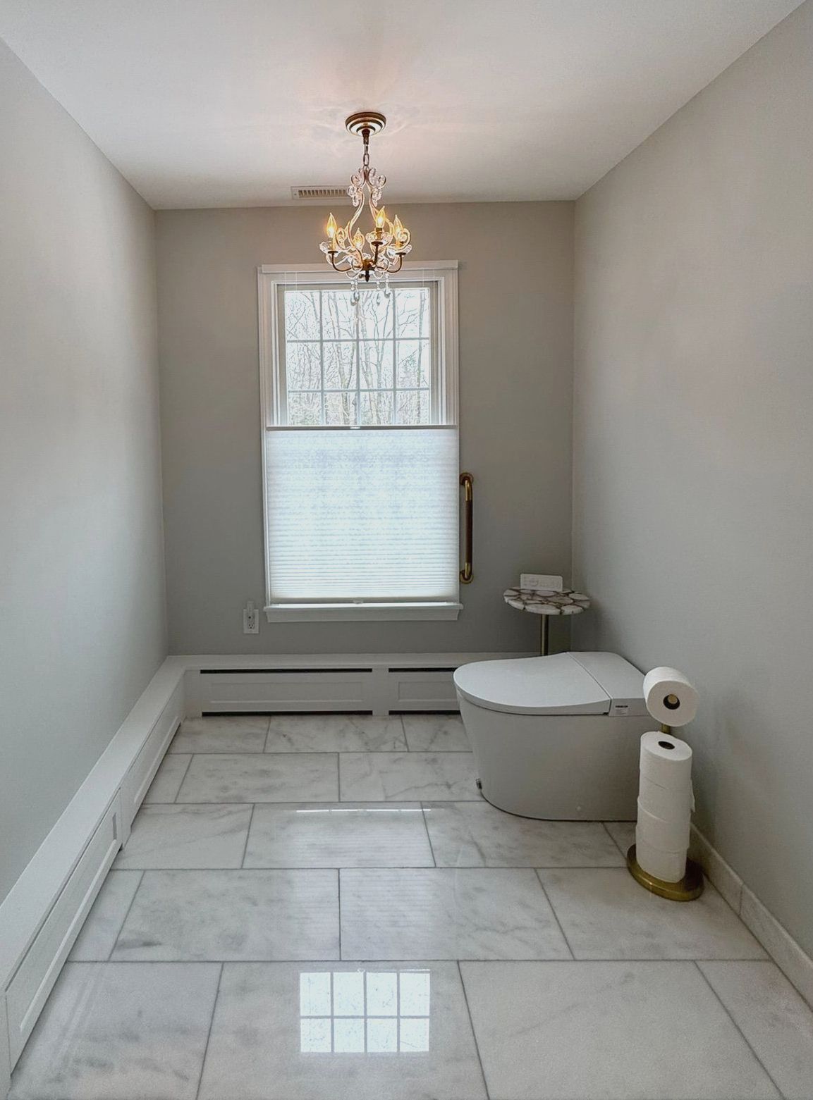 Empty modern bathroom with white tile floor, chandelier, window, and toilet paper holder.
