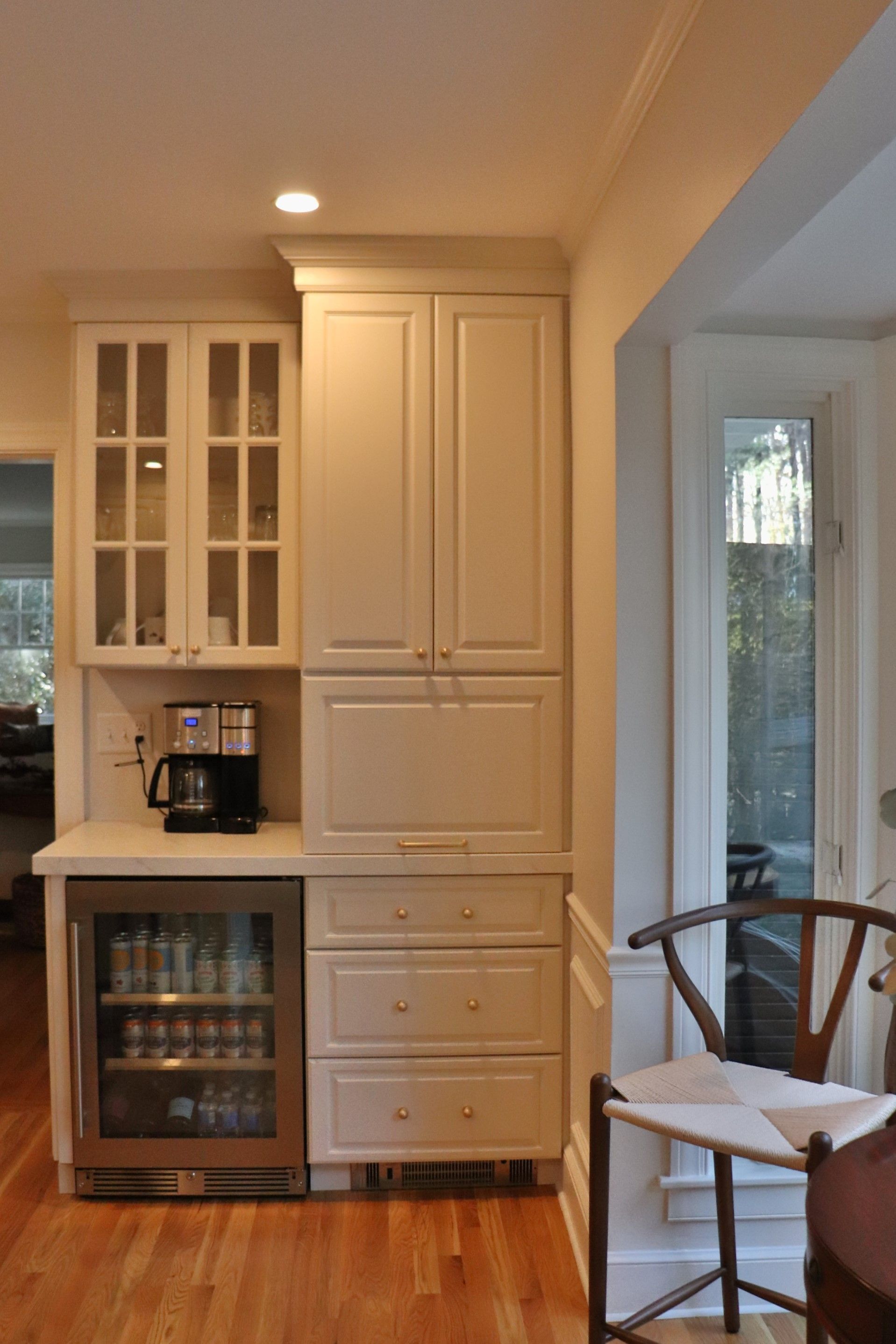 Built-in cream cabinets beside a small beverage fridge in a sunlit kitchen nook.