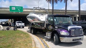 Tow truck hauling a white speedboat on a highway ramp under an overpass, with a green road sign nearby