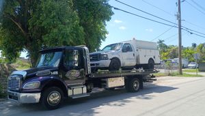 Tow truck carrying a white pickup on a sunny street beside trees and power lines