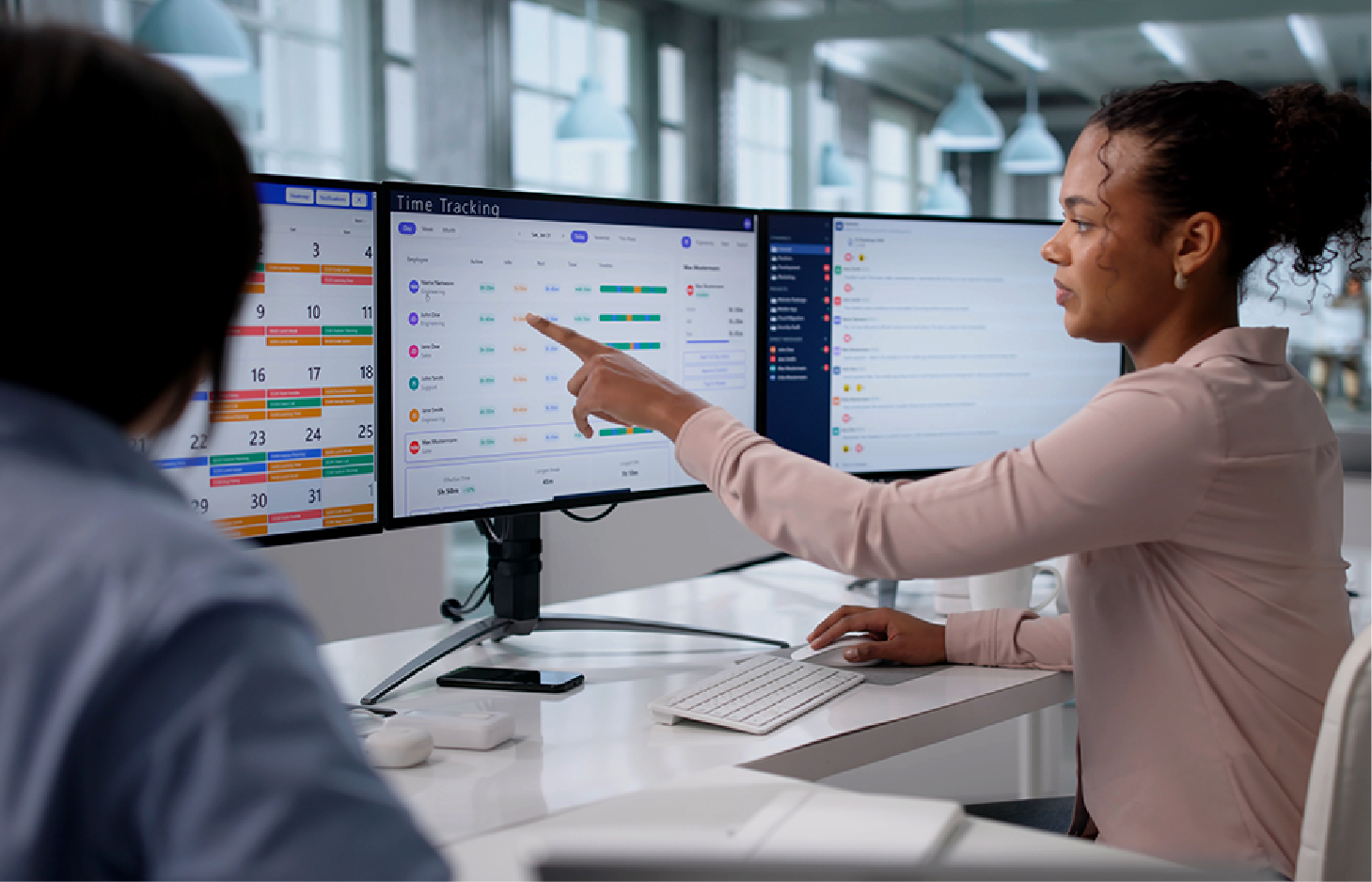 Two colleagues collaborate in a modern office, pointing at data on multiple computer monitors.
