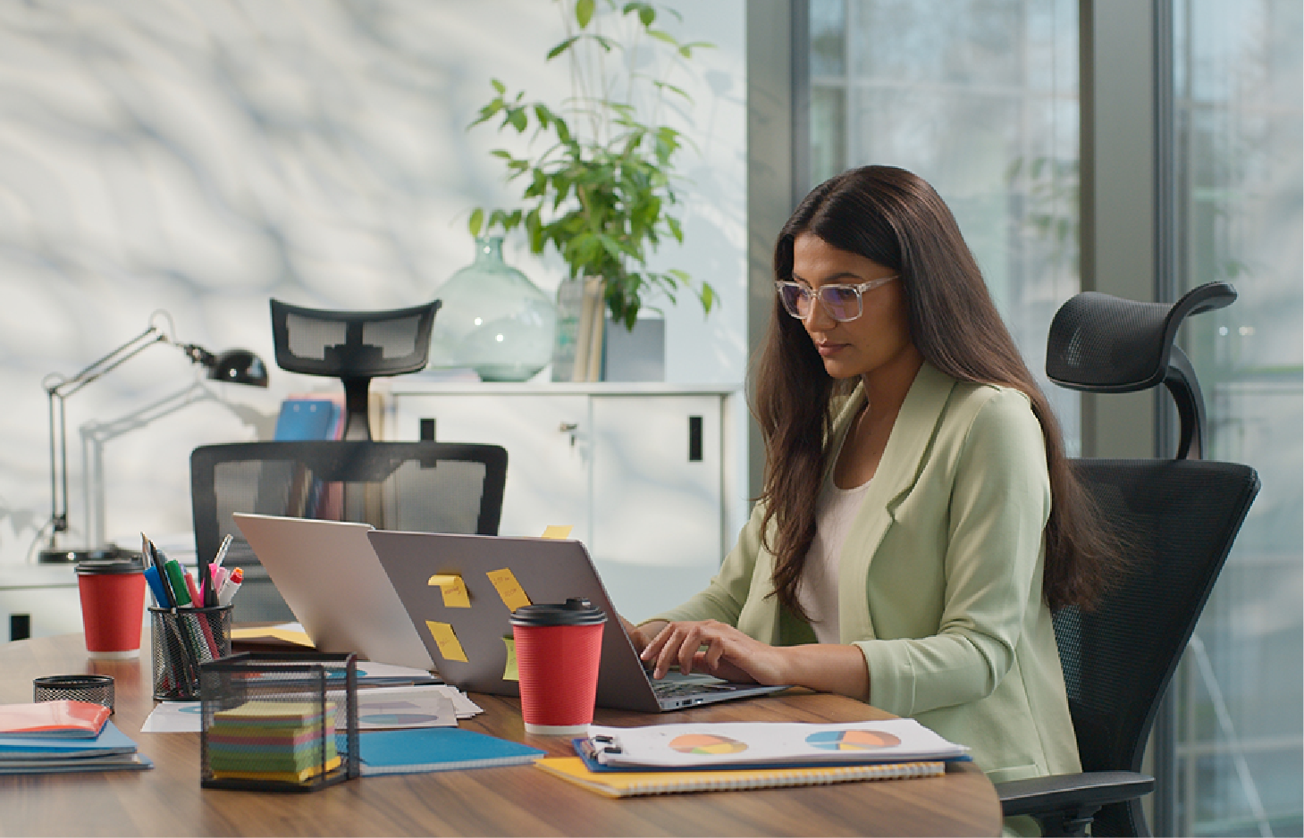 A person in a light green blazer works on a laptop at a wooden desk with office supplies and a coffee cup in an office.