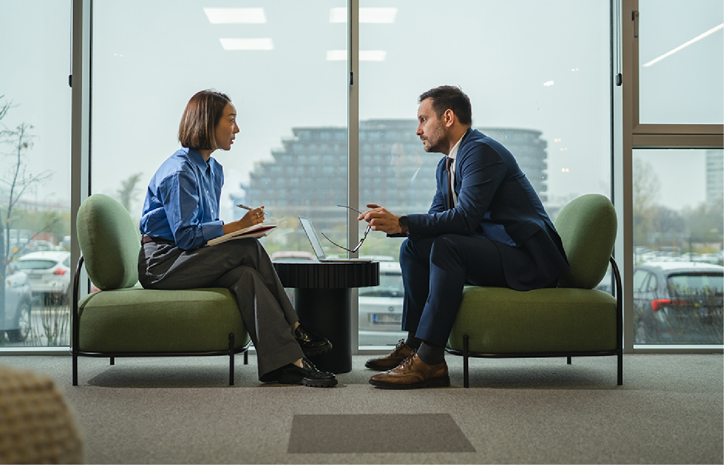 A person in a blue shirt and another in a suit sit on green chairs facing each other, conversing in a bright office.