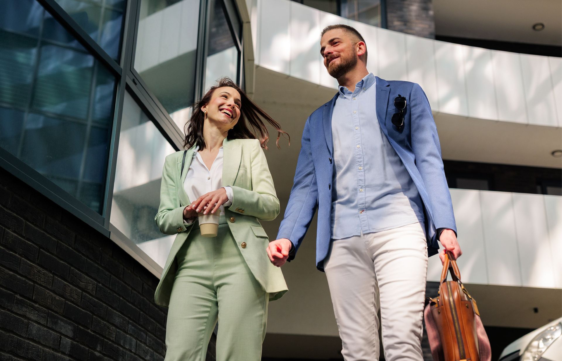 A smiling person in a light green suit walks alongside another person in a blue blazer carrying a briefcase.
