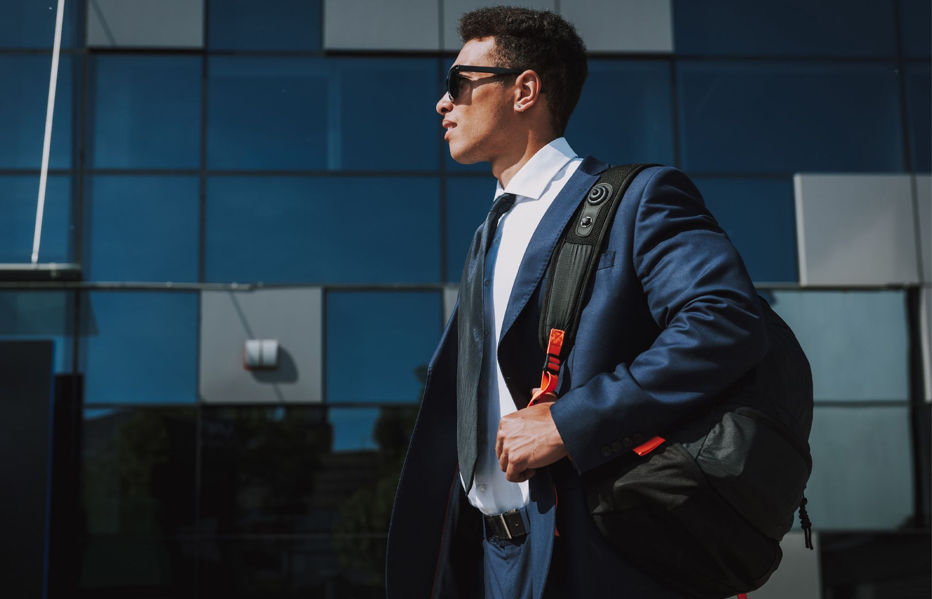 A person in a suit and sunglasses carries a backpack in front of a modern, glass-paneled building.
