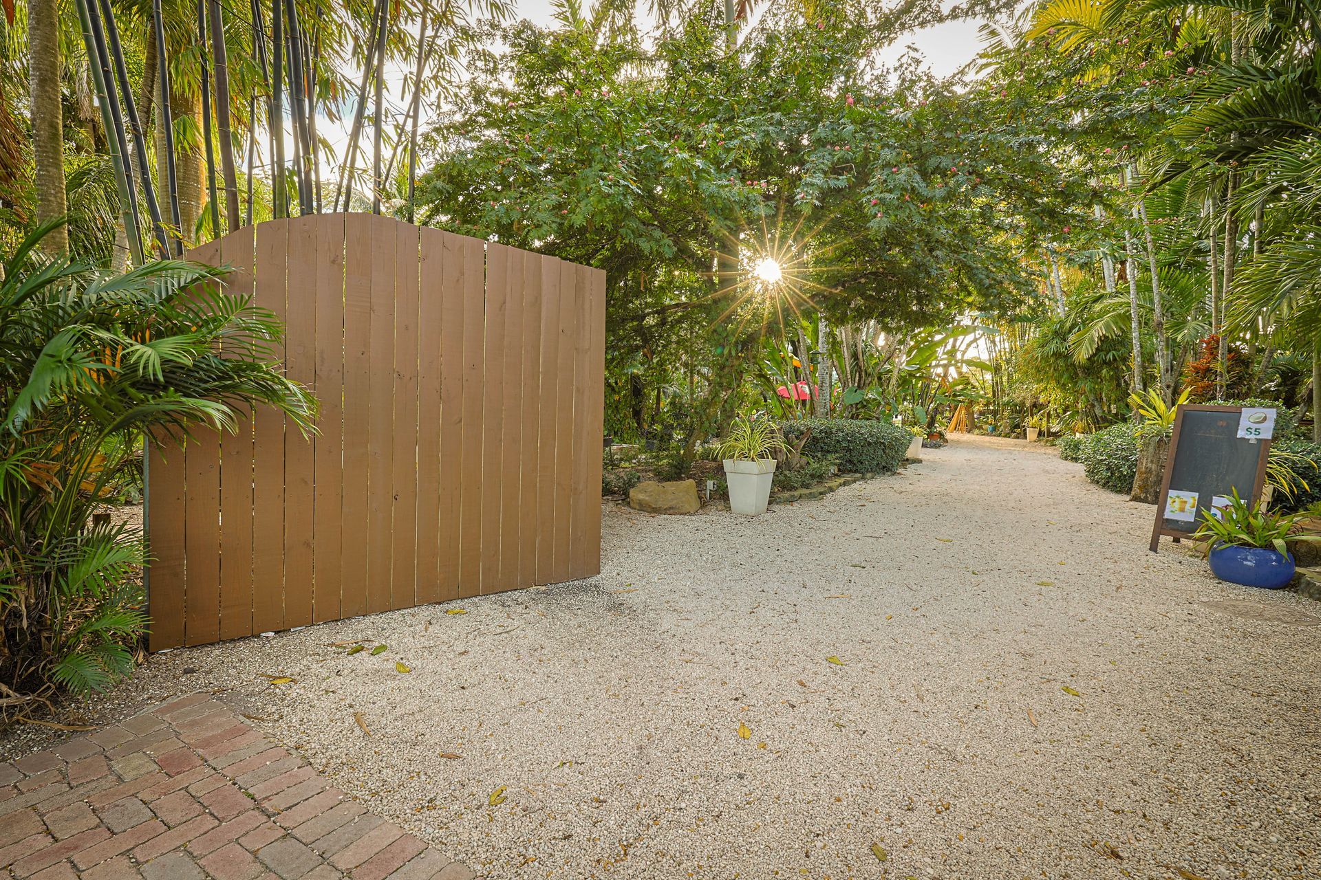 A gravel path leads through a lush, sunlit garden past a tall brown wooden privacy screen and a small chalkboard sign.