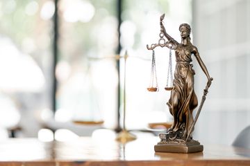 Statue of Lady Justice with scales and sword on a desk in front of open book and paper.