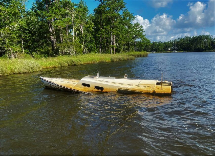 Small tan boat on a lake near a grassy shore and trees under a blue sky