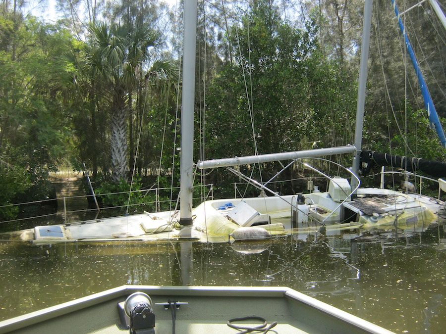 Capsized sailboat in calm water beside a wooded shore, with mast and rigging visible.
