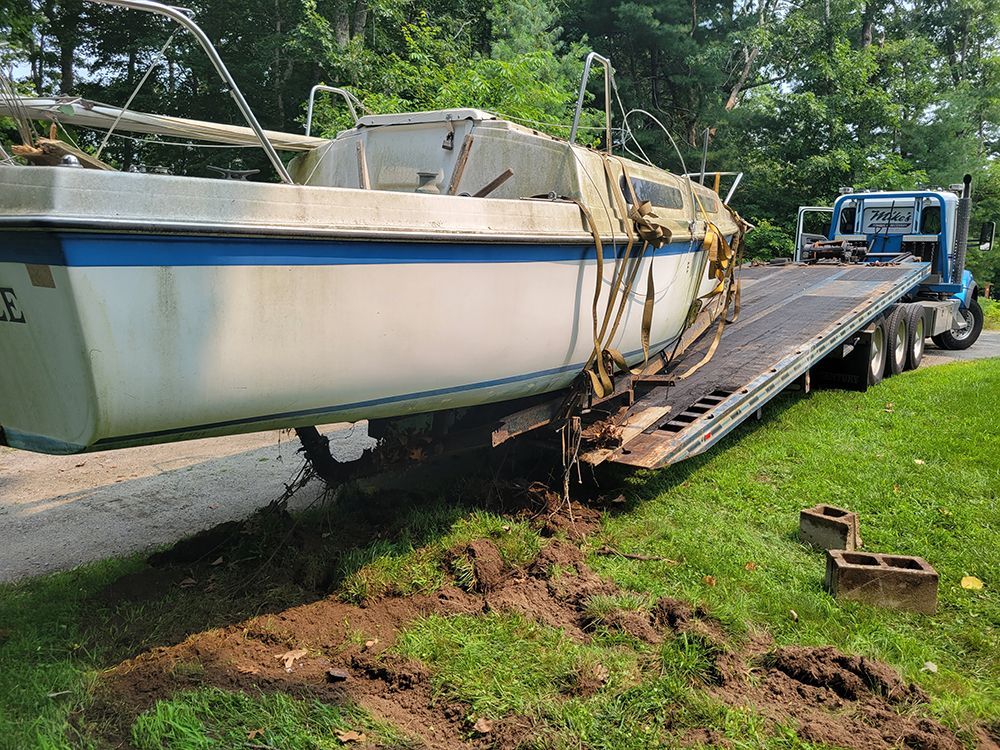 Large white sailboat on a trailer being hauled by a truck on a roadside, with straps and a ramp visible