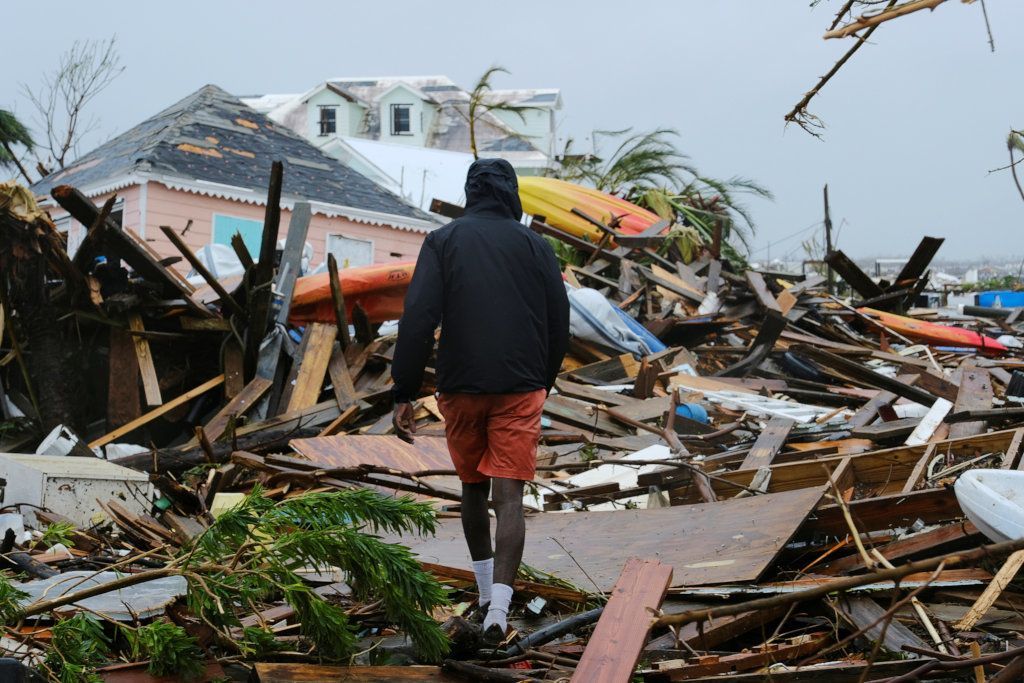 Person in yellow hat walks through storm debris beside a damaged house under gray skies