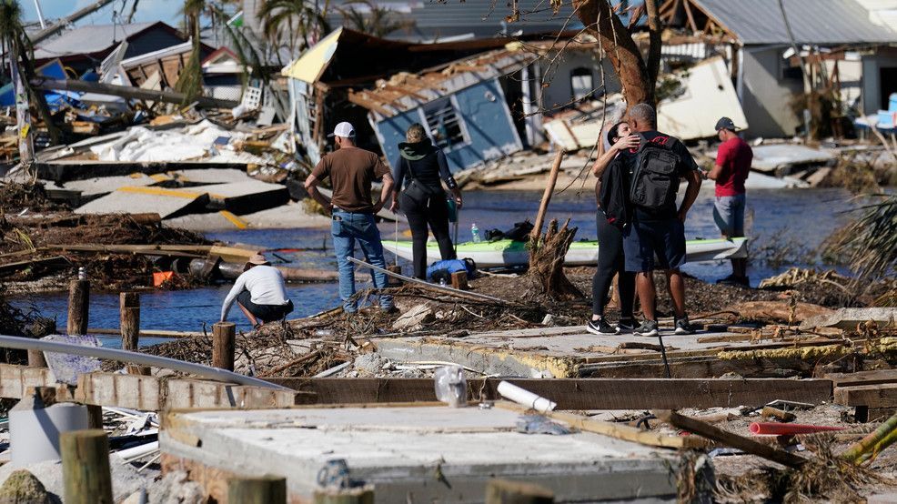 People walking through storm debris and flooded ruins, with collapsed houses in the background.