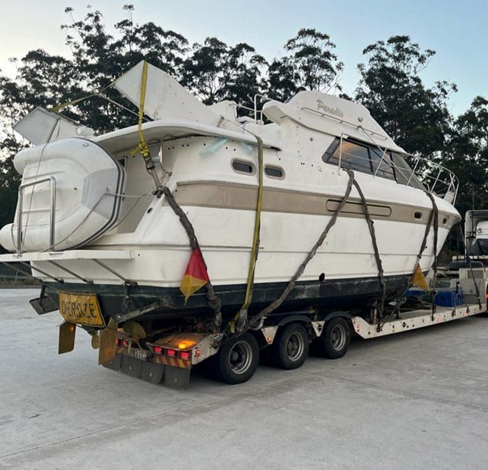 White yacht secured on a trailer with straps, being transported on a road near trees at dusk