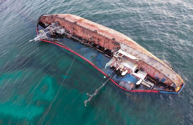 Aerial view of a rusted shipwreck partially submerged in turquoise water with a red boom surrounding it.