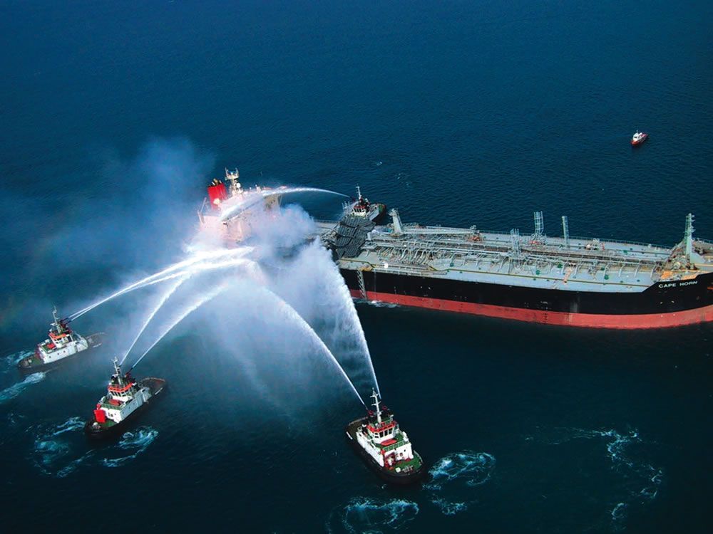 Fireboats spray water onto a large ship at sea, with smoke rising from the bow.