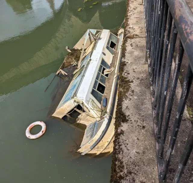 Partially submerged small boat beside a concrete canal wall, with a lifebuoy floating nearby