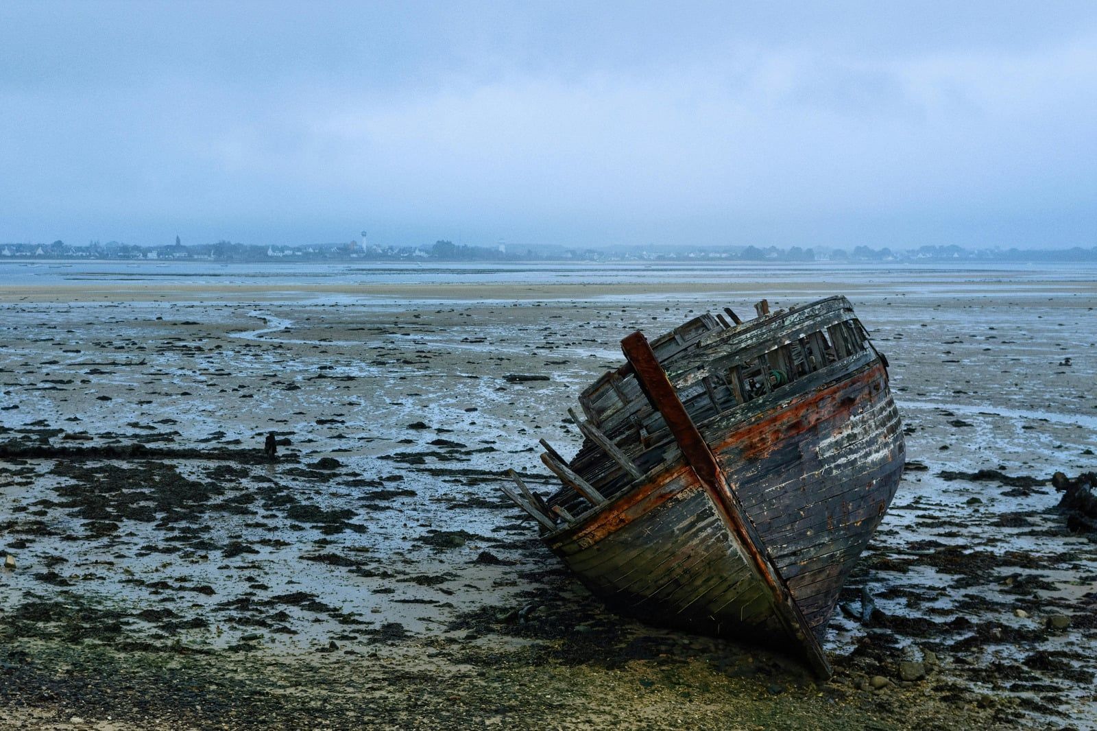 Stranded wooden boat on a muddy tidal flat under a gray sky, with a distant shoreline.